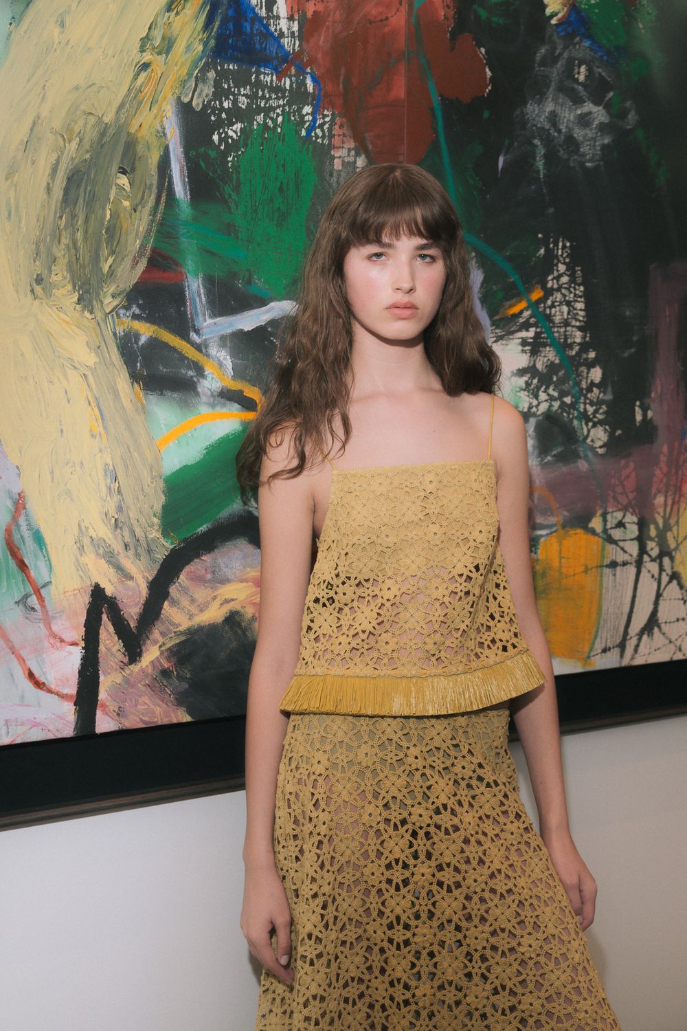Young woman in yellow crochet top and skirt poses before a colorful abstract painting.