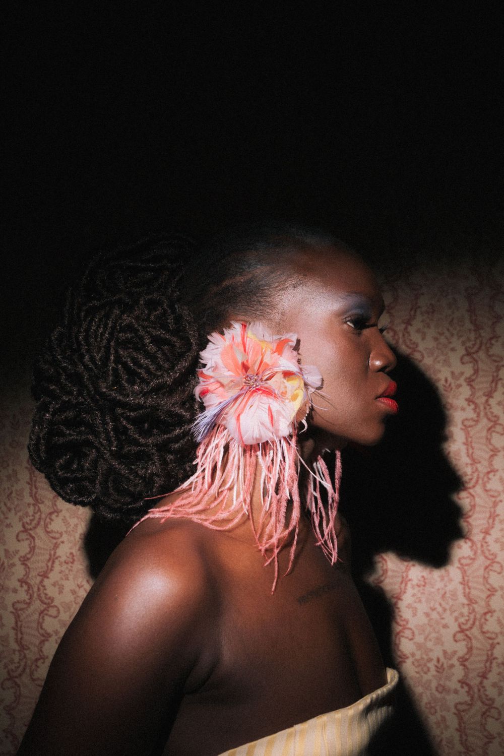 A Black woman with braided locs adorned with a pink flower hair accessory poses in dramatic side lighting.