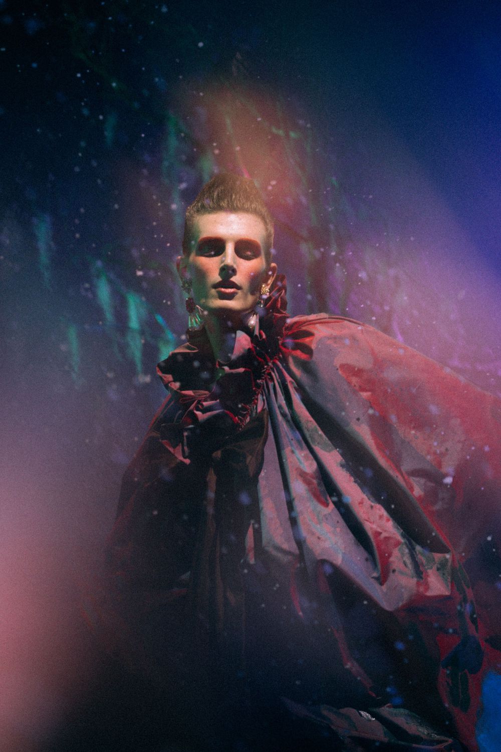 A dramatic figure in a dark red cape poses under colorful lights with a moody, ethereal atmosphere.