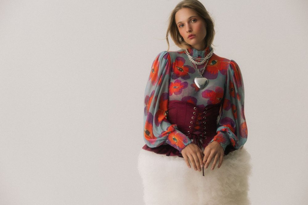 Woman wearing a floral blouse with puff sleeves, burgundy corset, and white fluffy skirt, seated against a gray background.