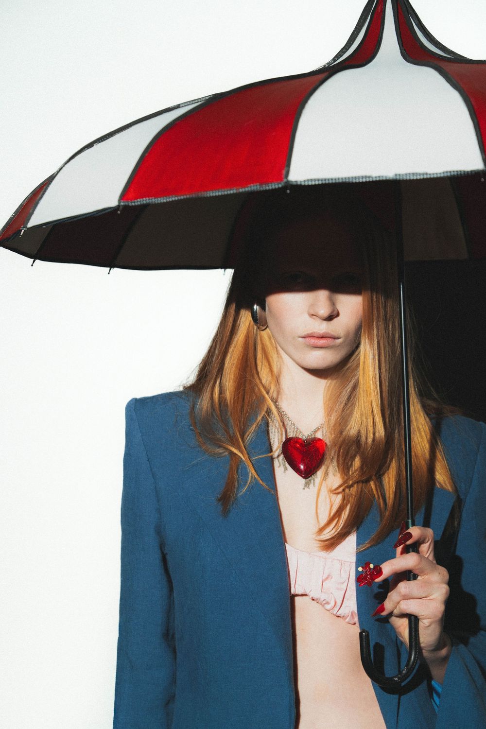Young woman in blue blazer holding red and white umbrella with heart necklace against white background.