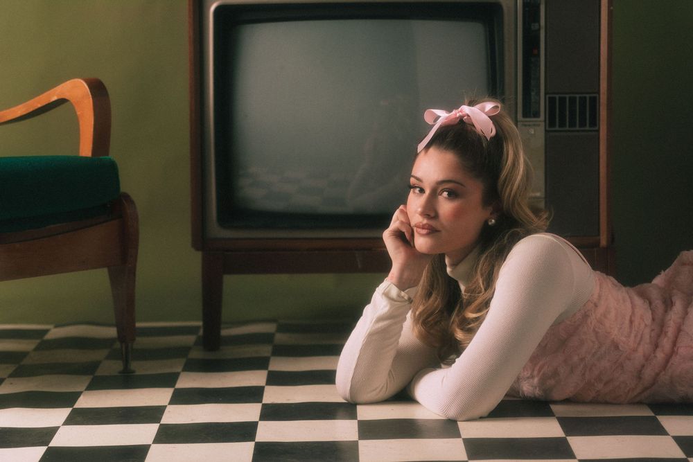 Young woman in pink bow and outfit lying on checkered floor beside vintage retro television in 1950s style setting.