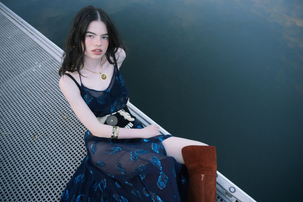 Young woman in blue floral dress and brown boots sitting on a dock over calm water.