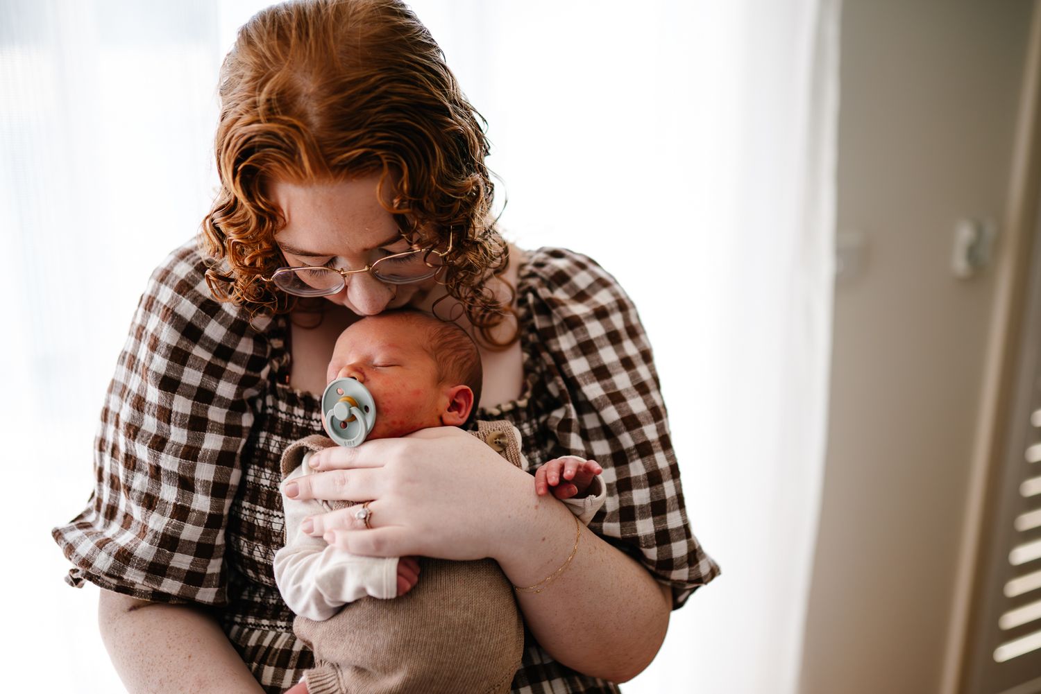 A mother in a checkered dress tenderly holds a sleepy newborn during a Melbourne newborn photography session at home.