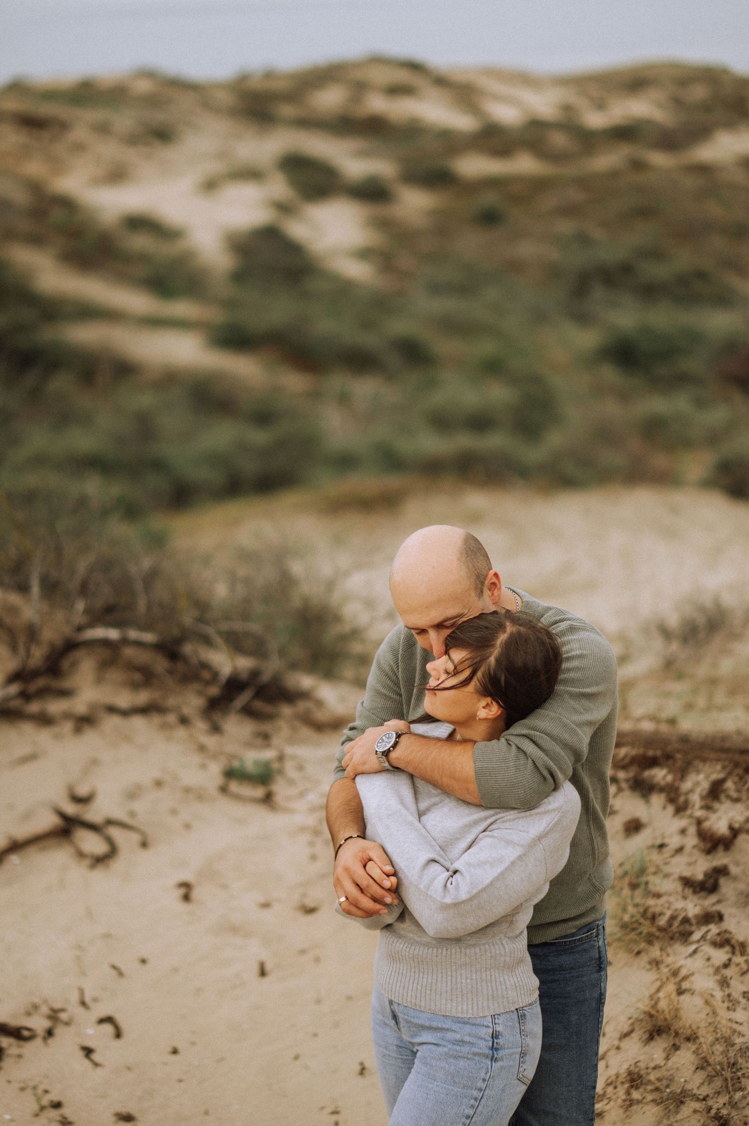 Couple embracing tenderly on sandy coastal dunes with green shrubs, a romantic outdoor engagement photo session by the sea, Netherlands