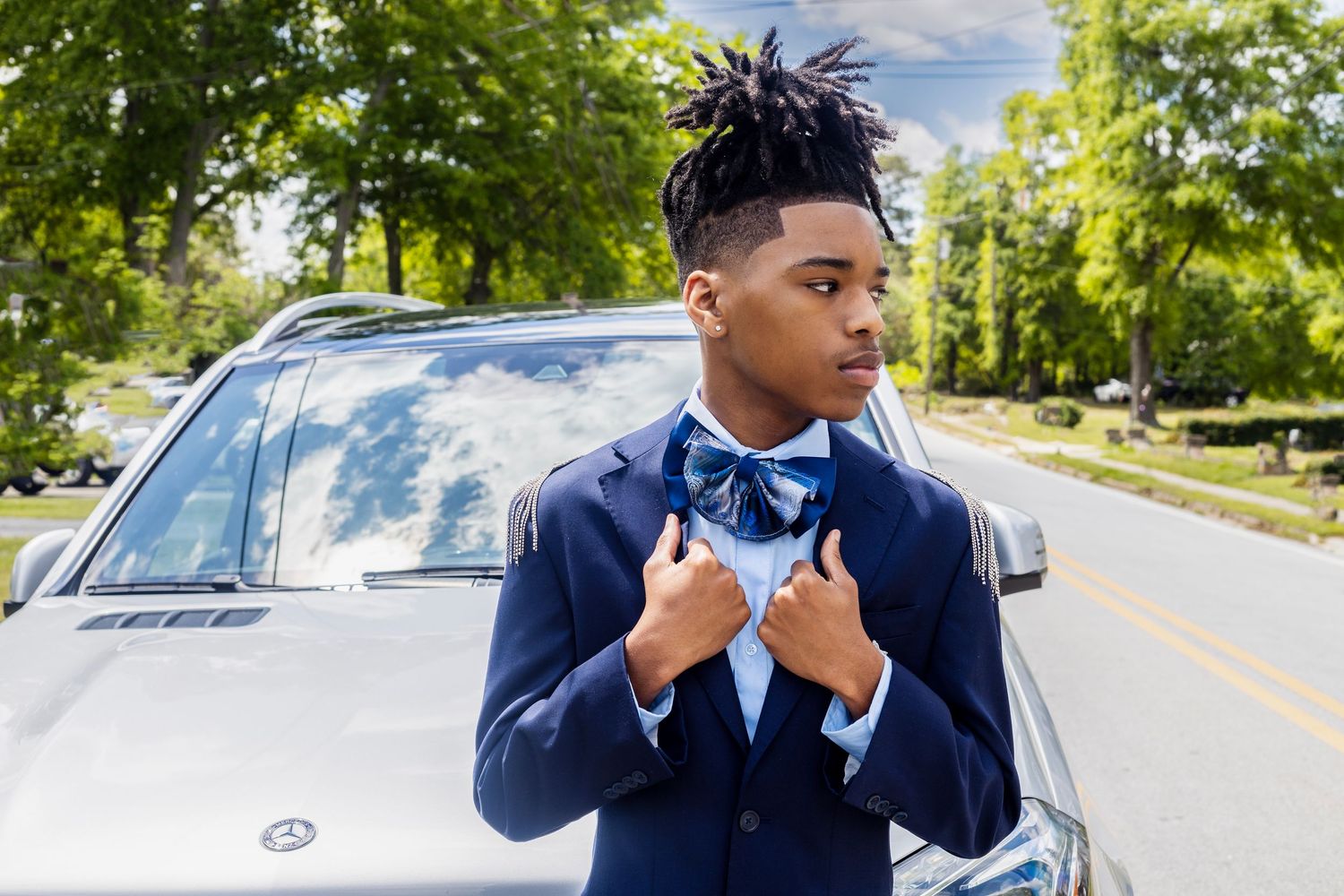 Young person in navy suit with bowtie poses confidently next to car.