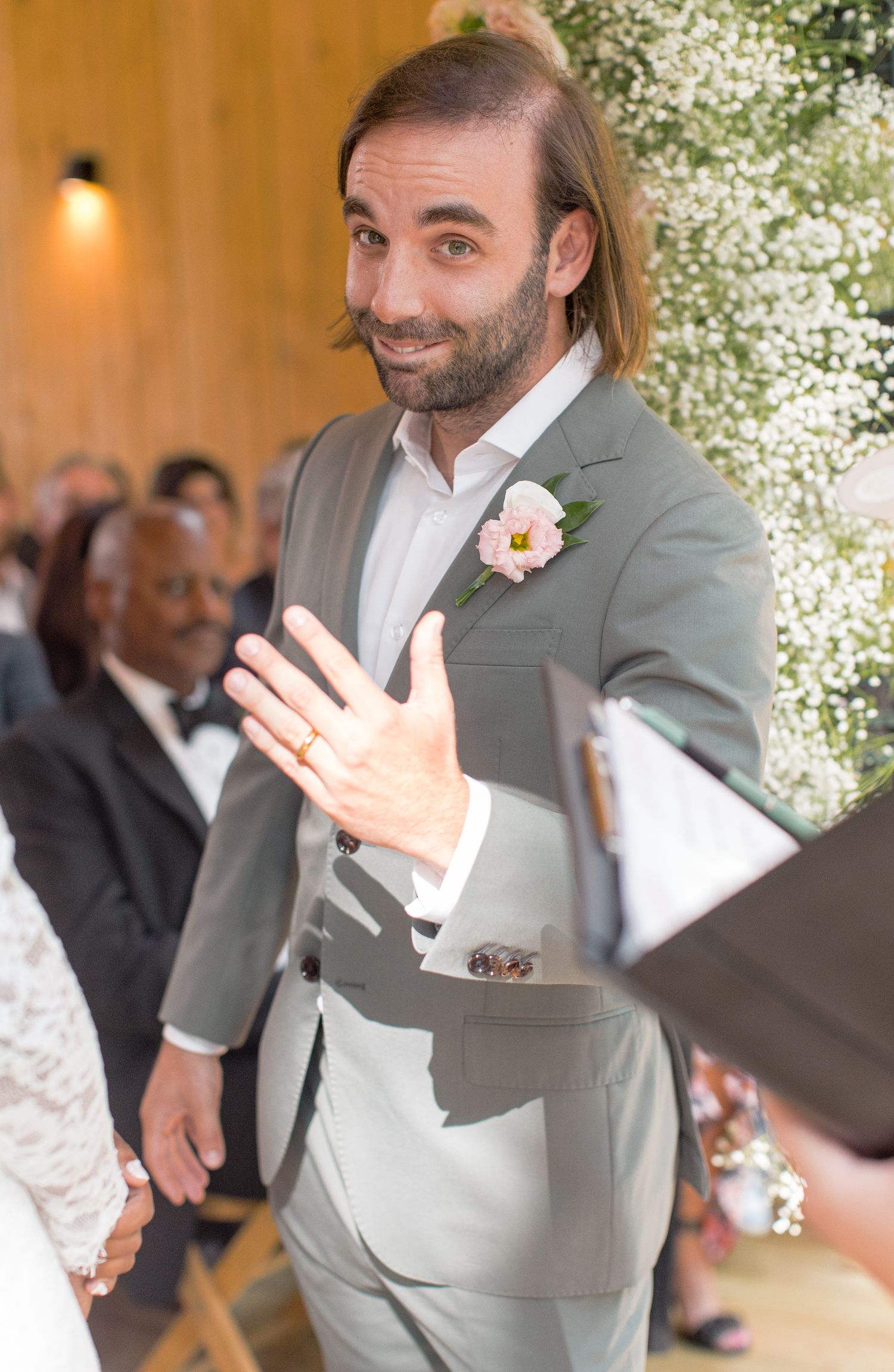 A person in a light gray suit and white shirt gestures during what appears to be a wedding reception.