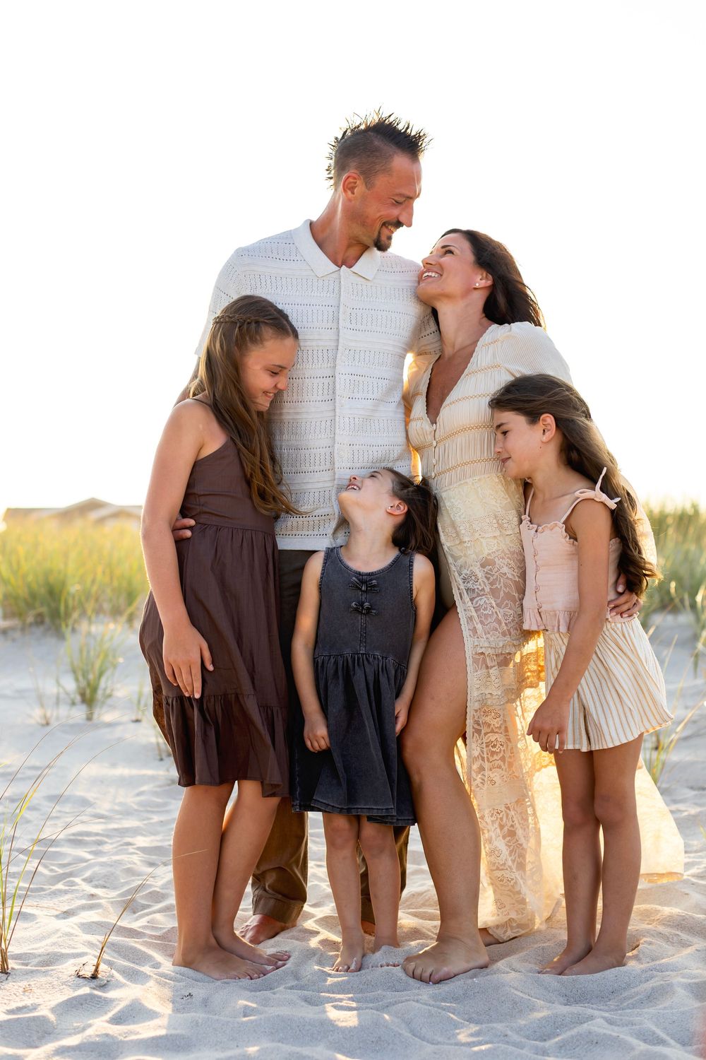 Family poses for candid beach photos at sunset while sitting in sand and sharing joyful moments together.