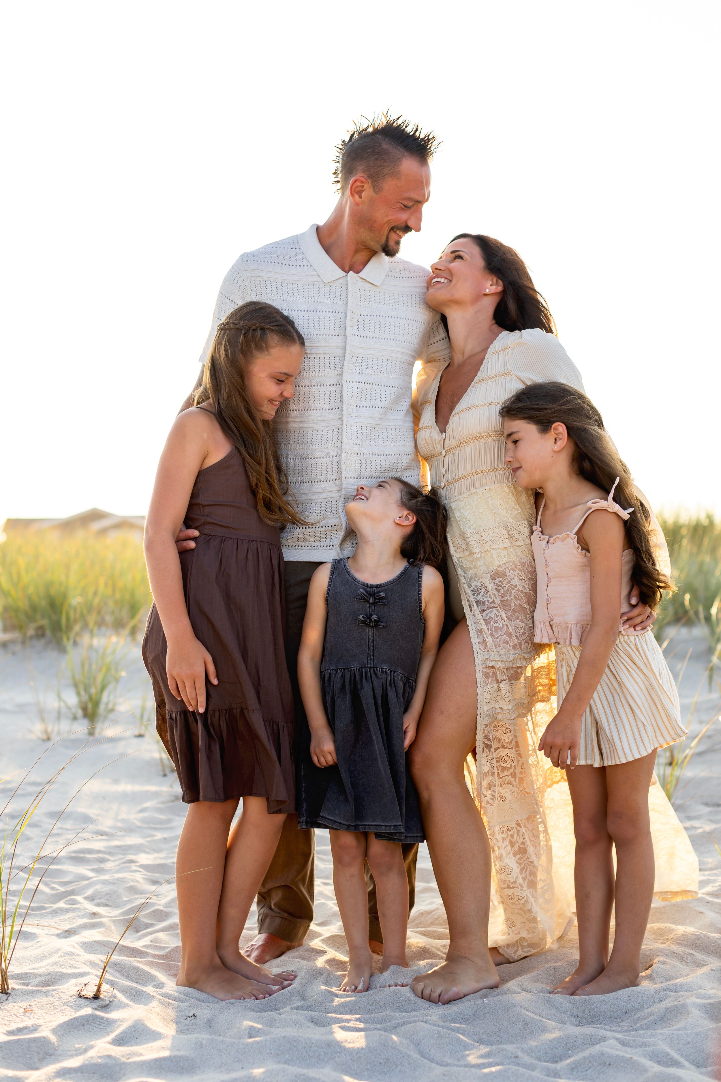 Family poses for candid beach photos at sunset while sitting in sand and sharing joyful moments together.