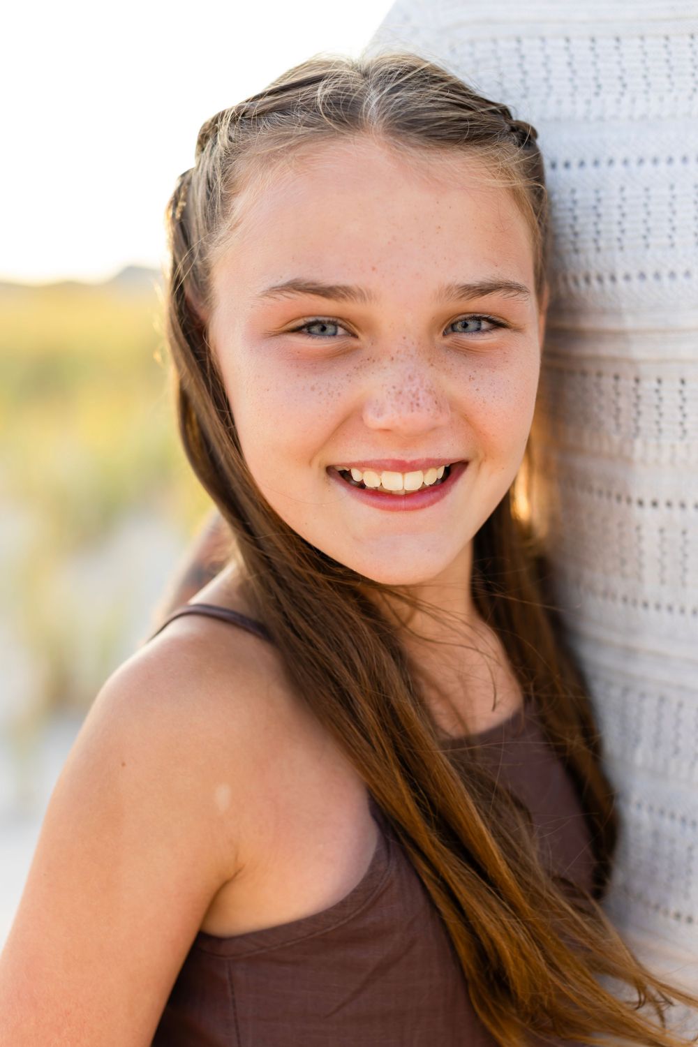 A close-up outdoor portrait shows a bright smile against a white wall with sunlit scenery in the background.