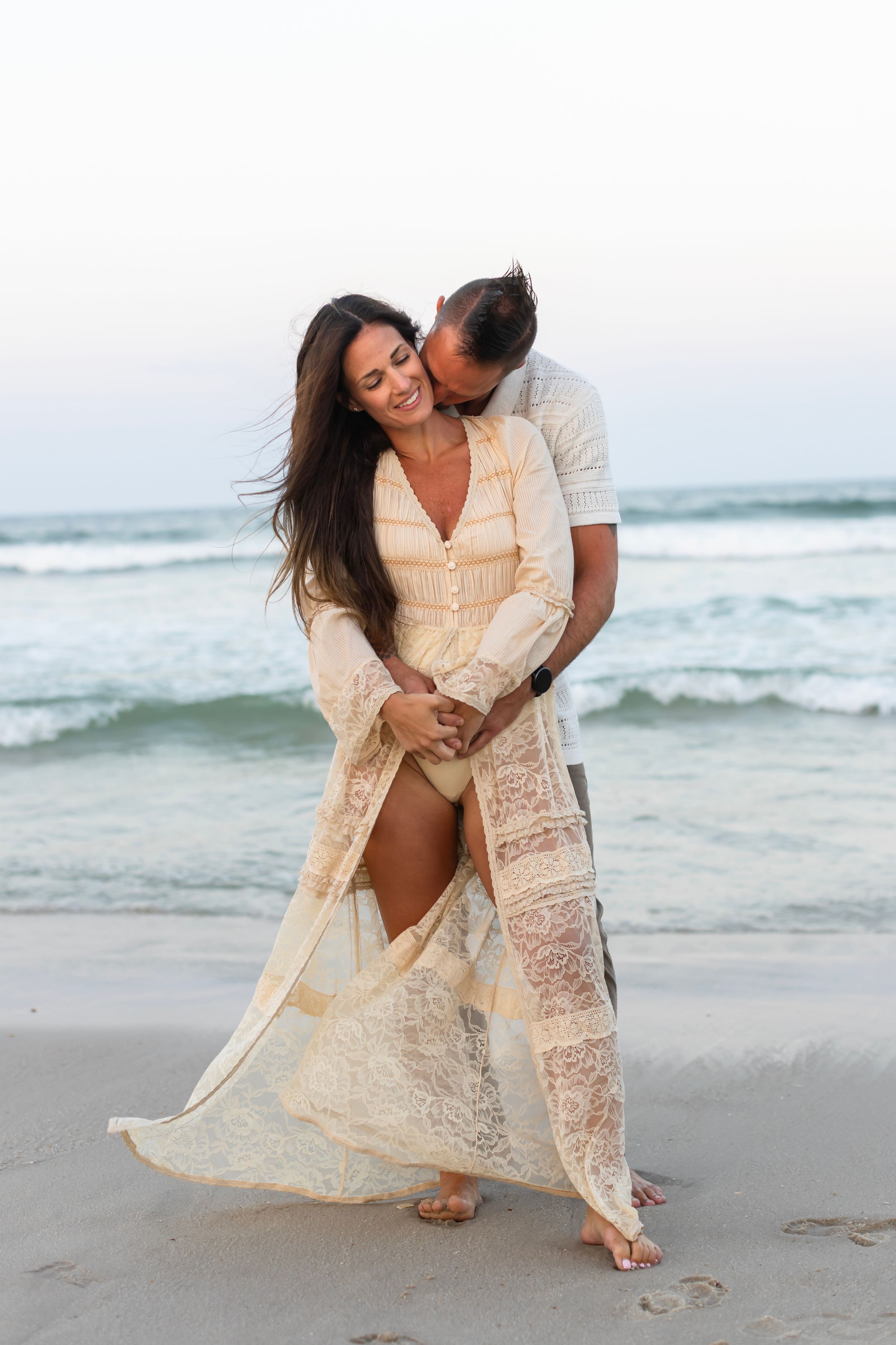 A romantic couple embraces on a sandy beach with crashing waves as the sun sets in the background creating a dreamy mood.