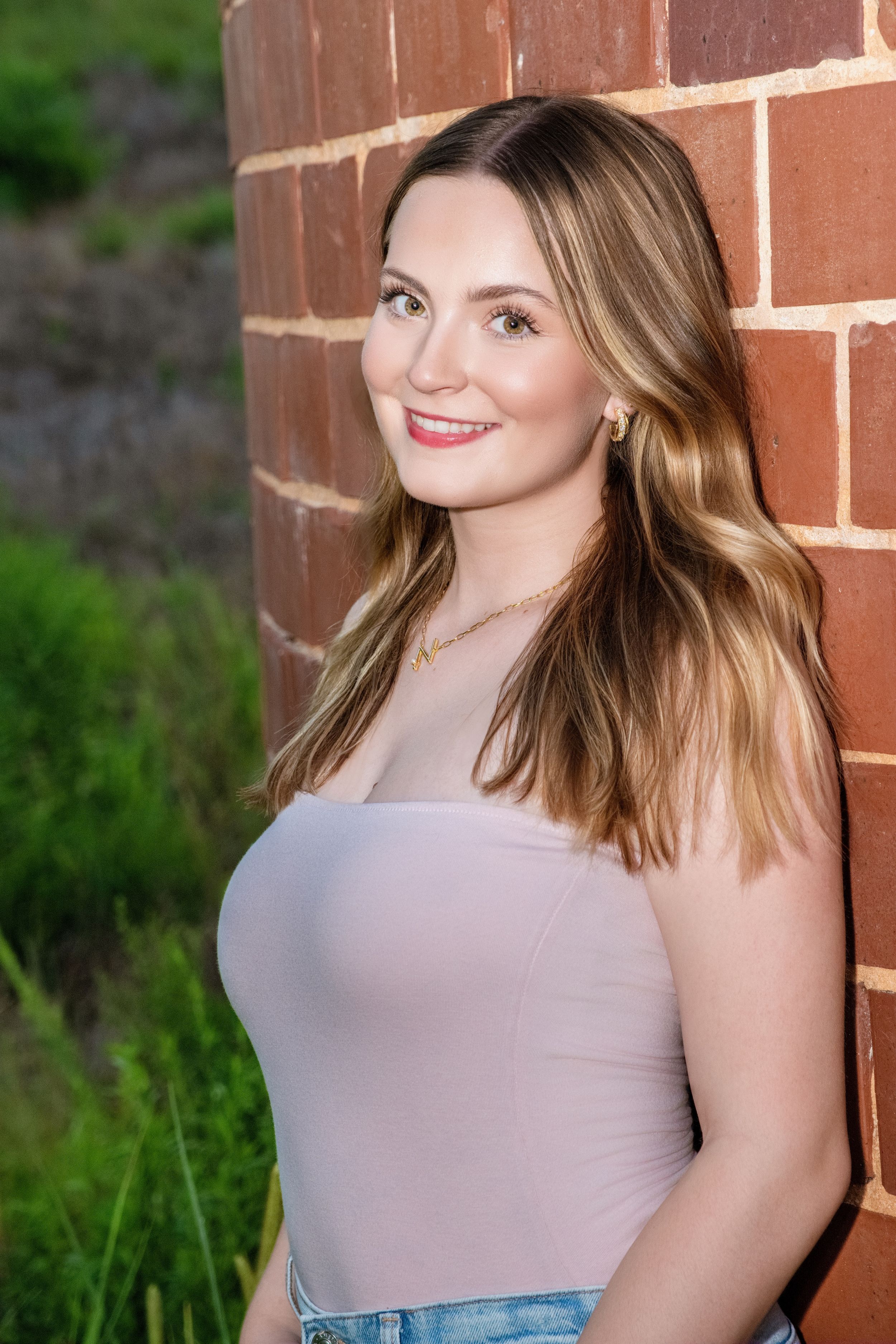 A person wearing a gray tank top poses against a red brick wall with green foliage in the background.