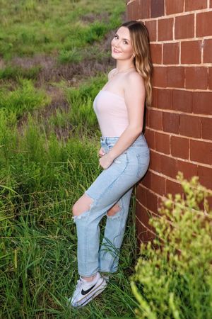 A person in a pink top and jeans poses casually against a brick wall outdoors surrounded by green grass and foliage.