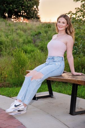 A young figure wearing ripped jeans sits casually on a wooden bench in a park setting during golden hour.