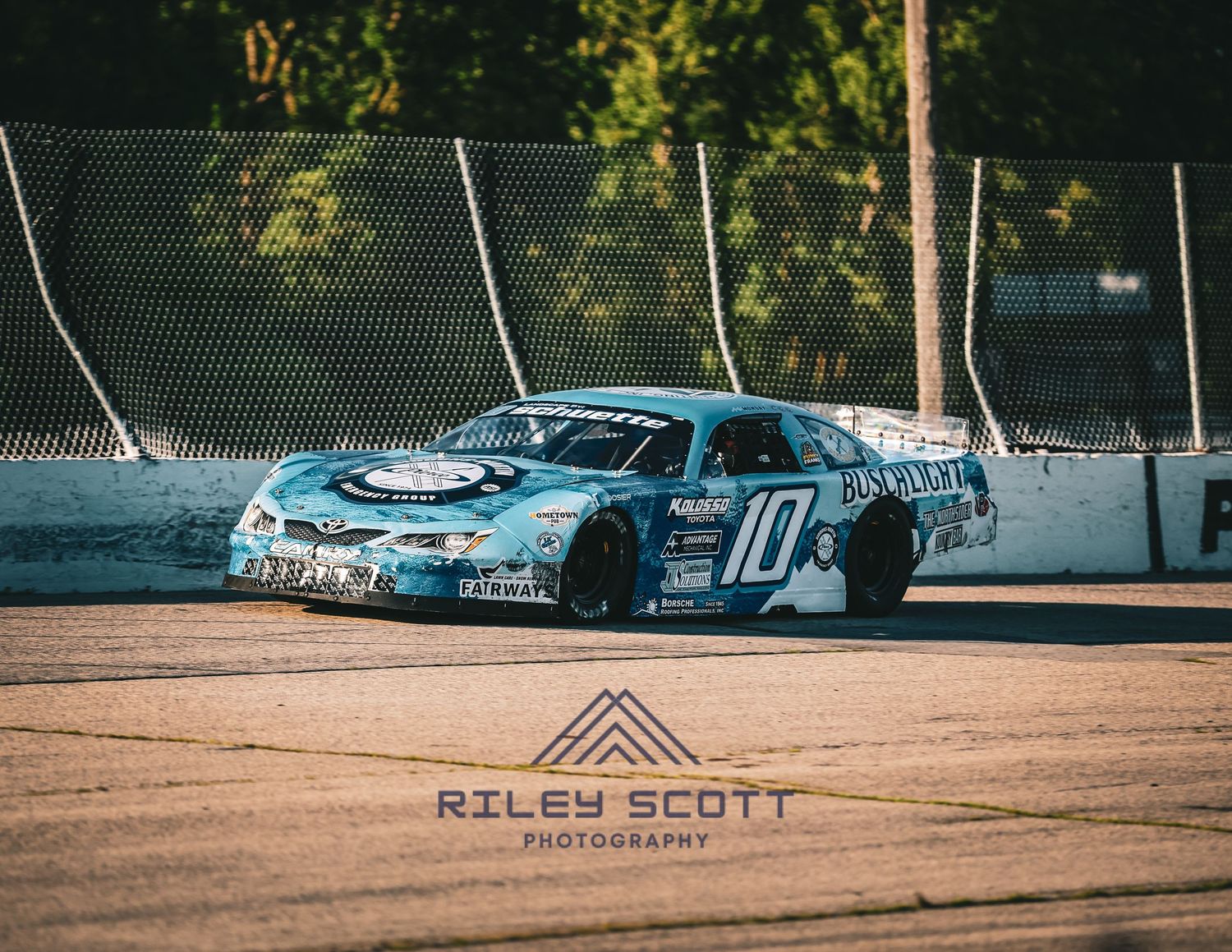 A blue drift car with custom graphics sits parked on a dirt surface near a chain link fence.