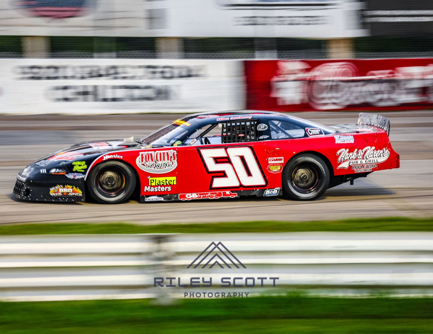 A race car drives at high speed along a racetrack, displaying prominent red and black sponsorship livery.