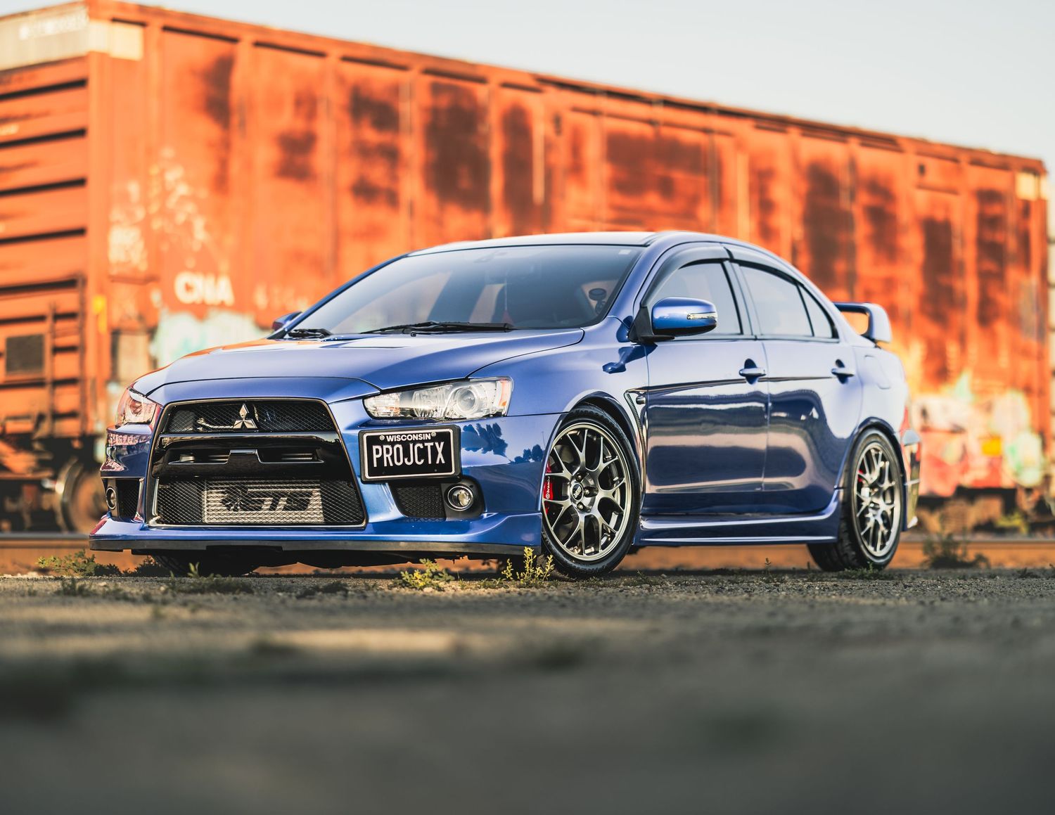 Blue Mitsubishi Lancer Evolution parked near red freight train cars, showcasing sporty body kit and alloy wheels.