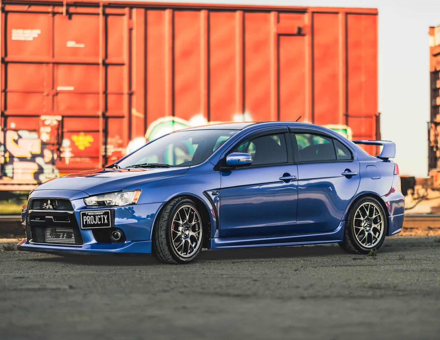 Blue Mitsubishi Lancer Evolution X sports sedan with rear spoiler and aftermarket wheels parked near red freight containers.