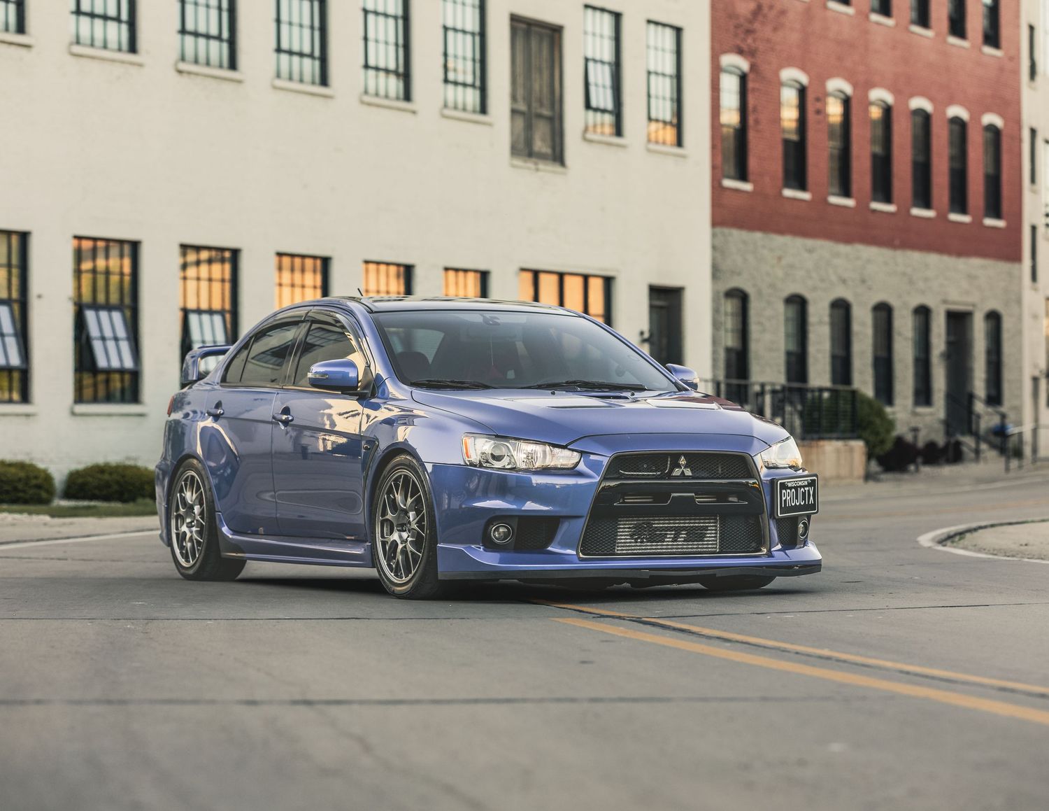 Blue Mitsubishi Lancer Evolution parked on urban street corner near brick building, showcasing sporty body kit and aggressive stance.