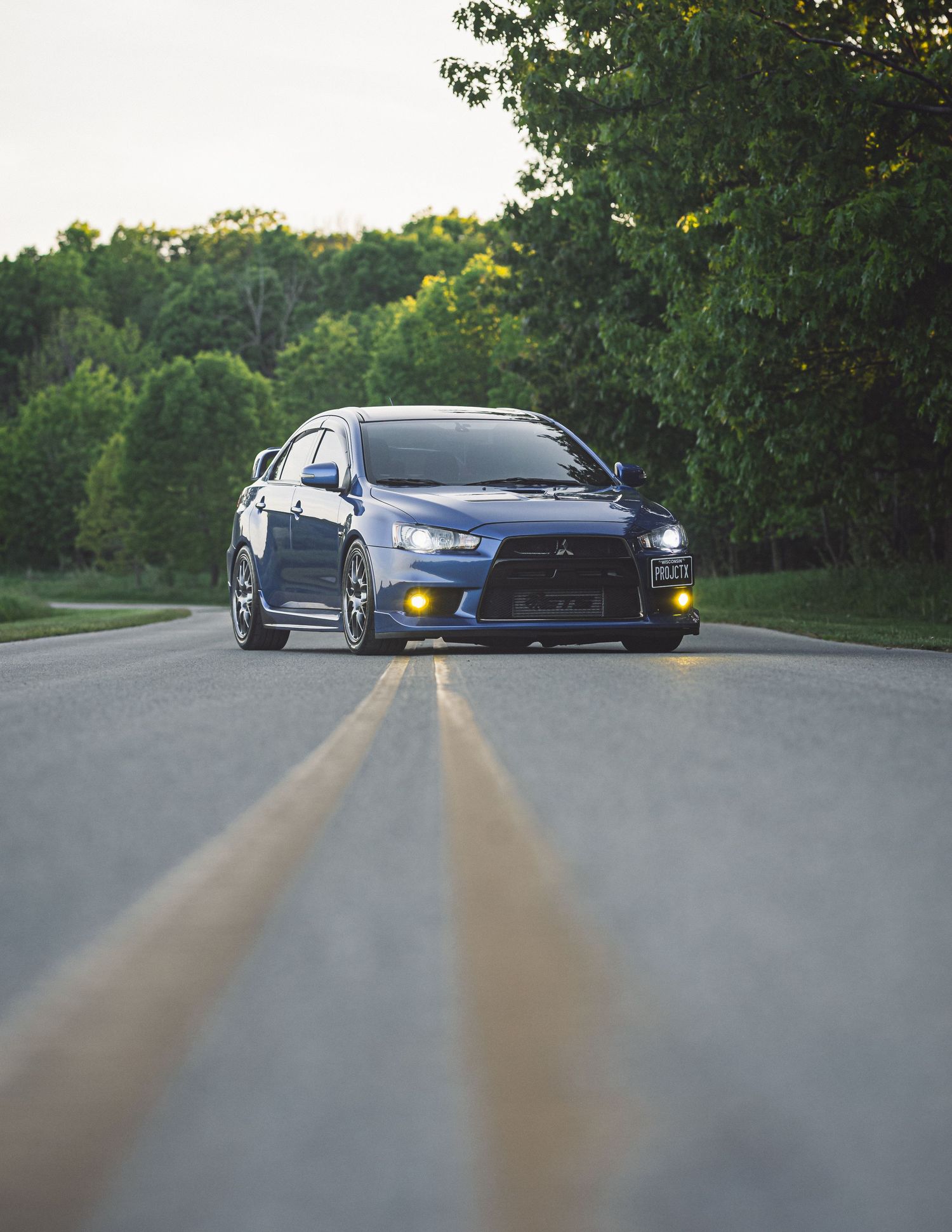 Blue Mitsubishi Lancer Evolution driving on a tree-lined road with yellow fog lights illuminated.