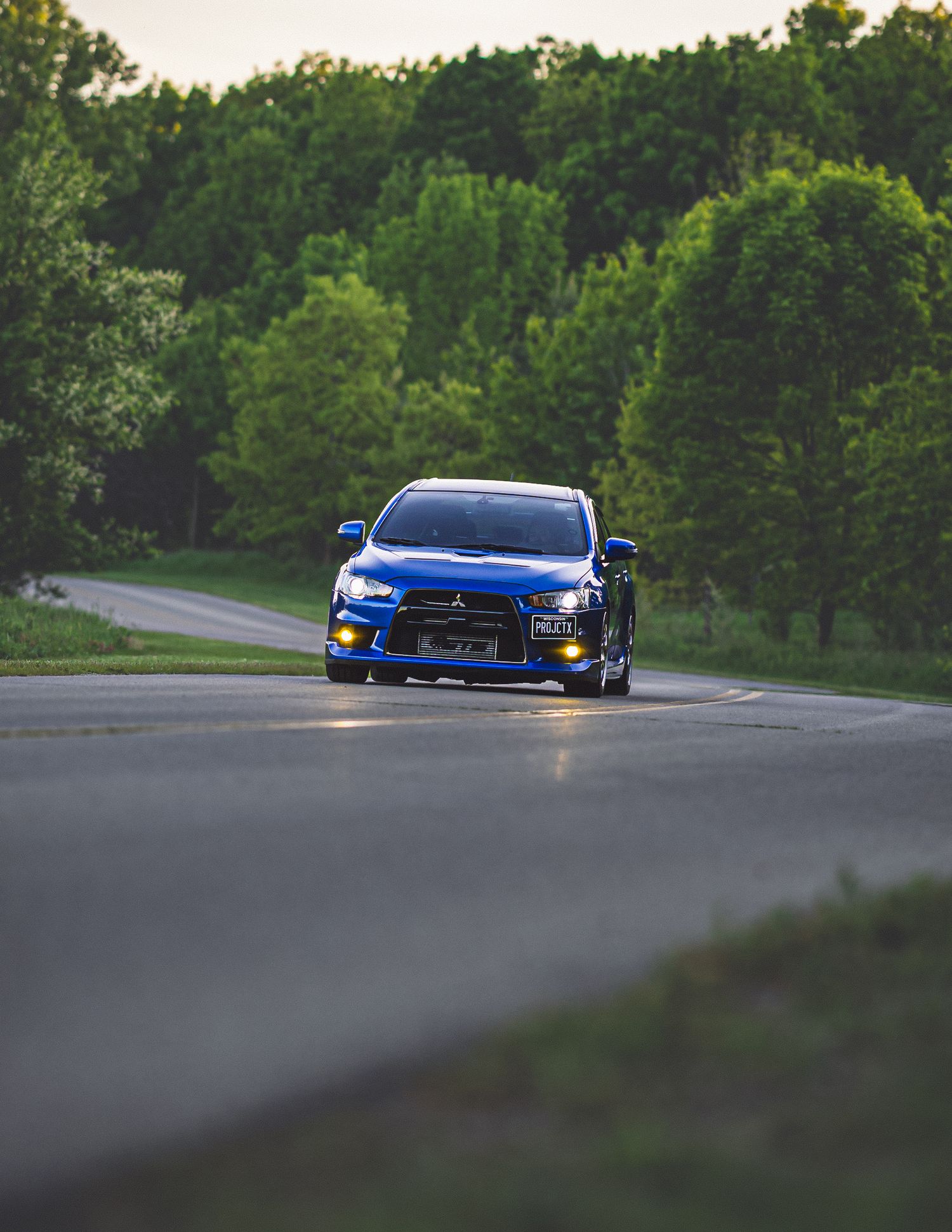 A blue Mitsubishi Lancer Evolution drives on a winding road surrounded by lush green trees at dusk.