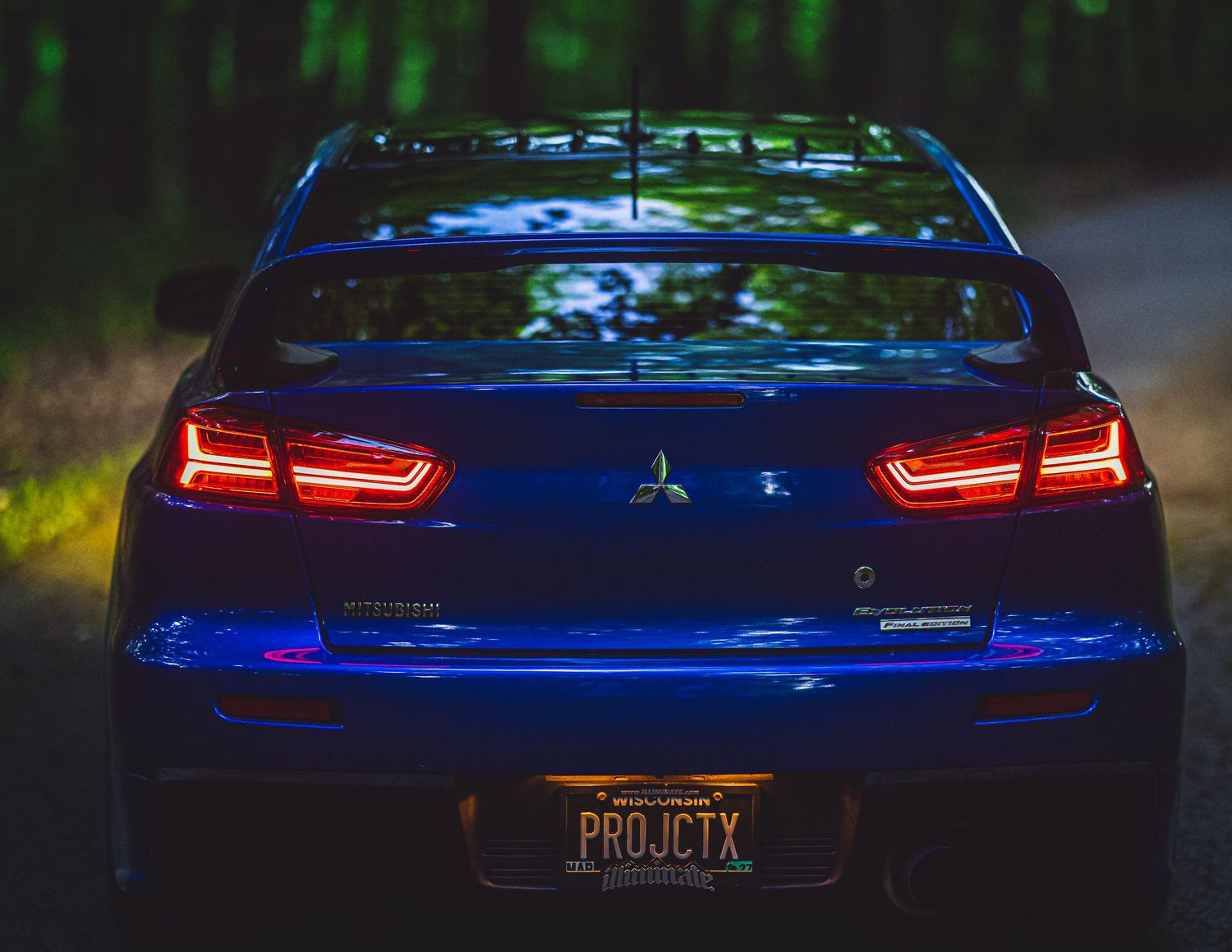 Rear view of a blue Mitsubishi car with red tail lights and custom PROJCTX license plate in a dark forest setting.