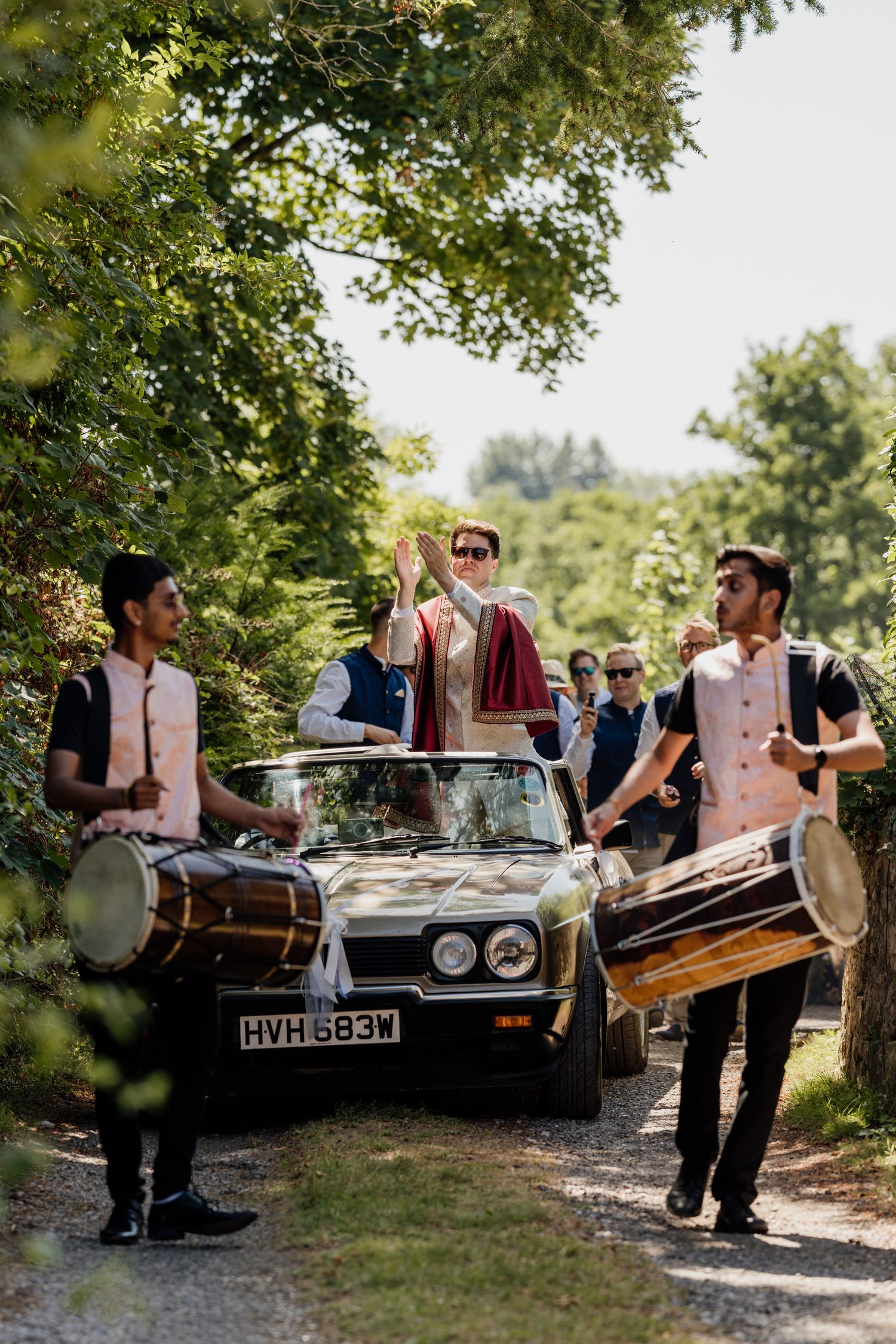 Wedding guests gather around classic vintage car in outdoor garden setting.