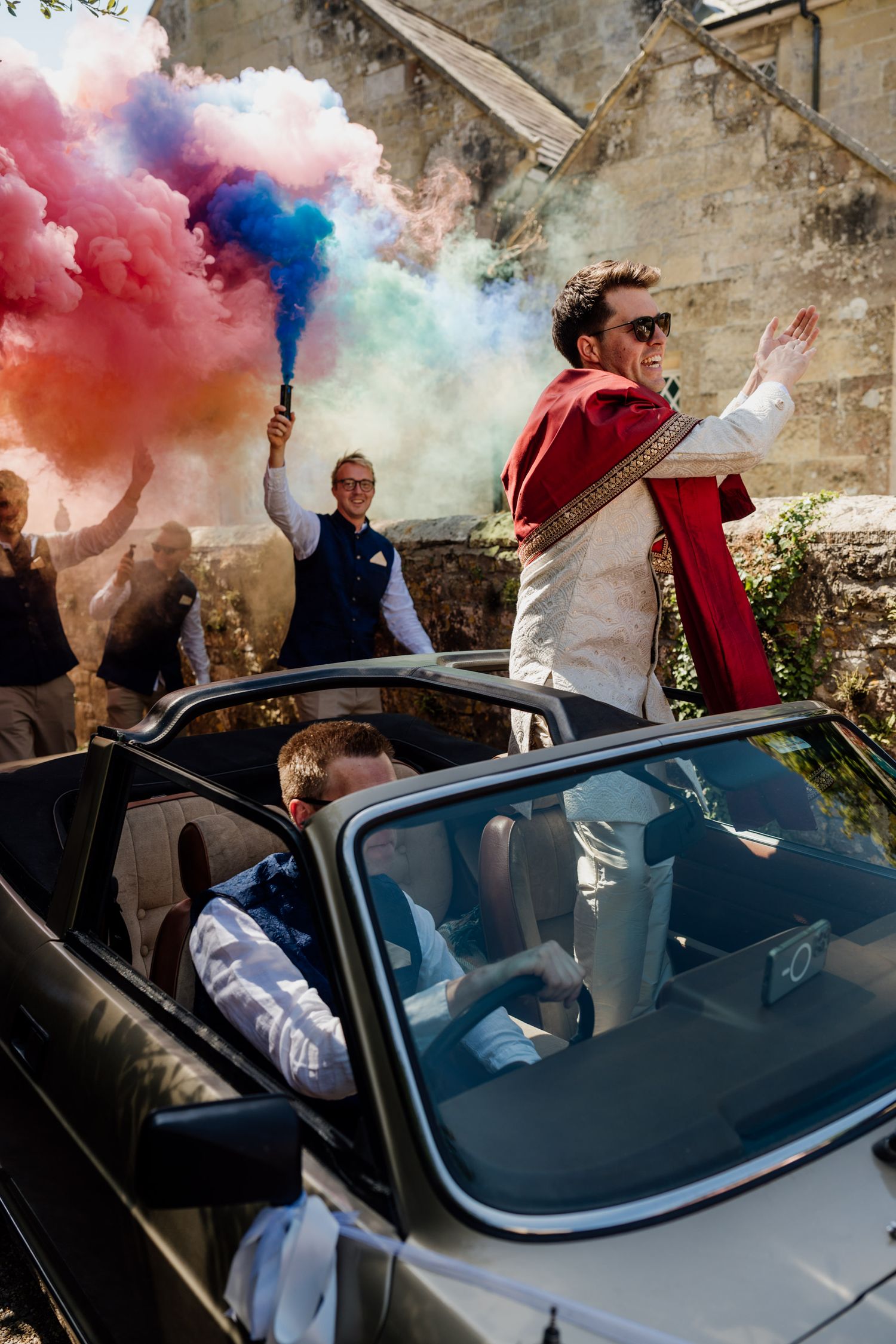 Wedding celebration with colorful smoke bombs creating vibrant atmosphere around a vintage car.
