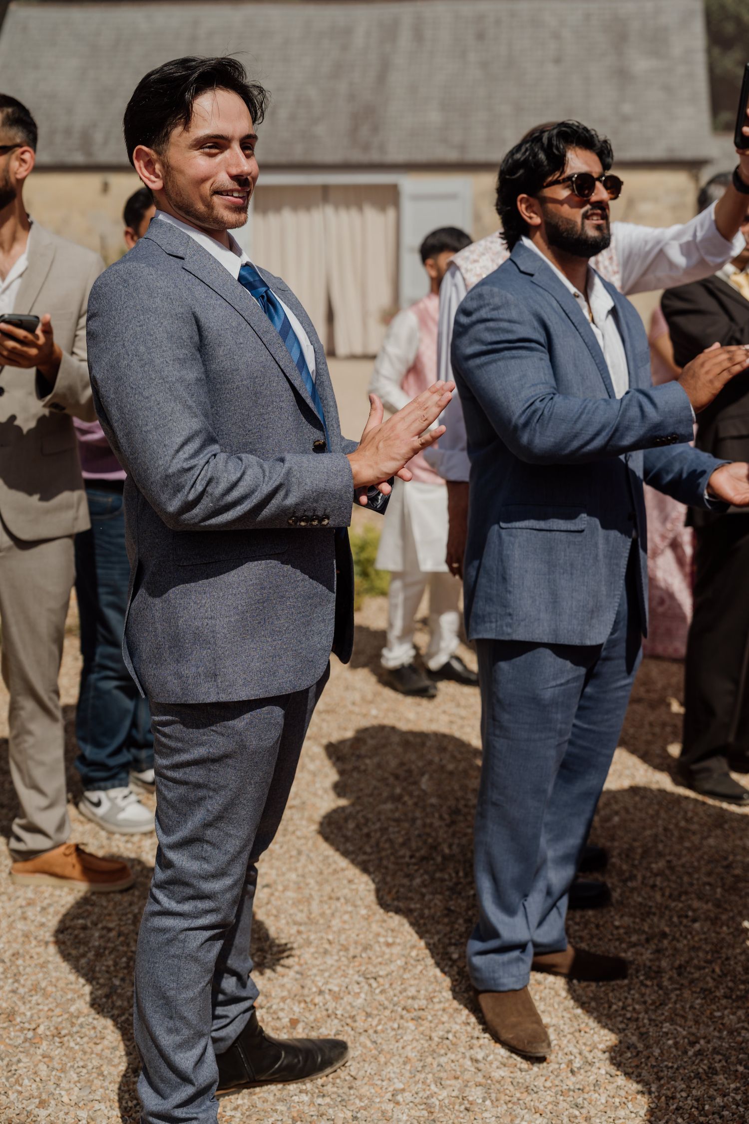 Wedding guests in grey suits stand together applauding and celebrating during an outdoor ceremony.