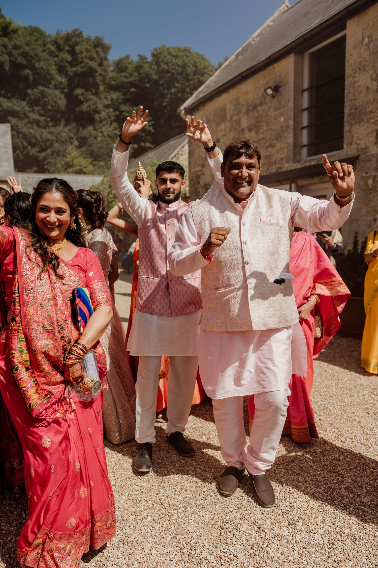 A festive celebration outdoors with people in traditional white and pink attire dancing against a stone building backdrop.