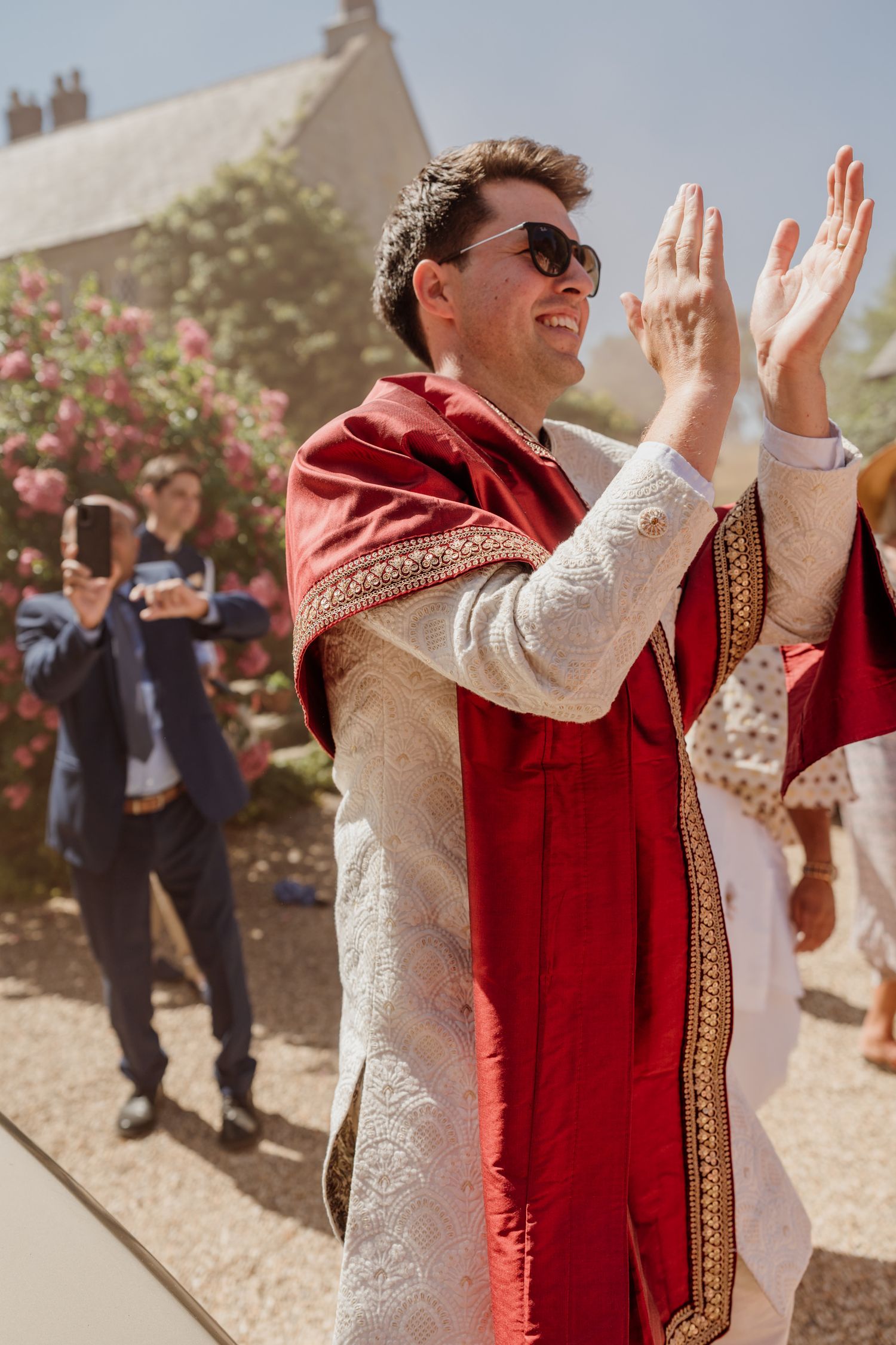 Wedding guest in white and red traditional attire claps and celebrates during outdoor celebration.