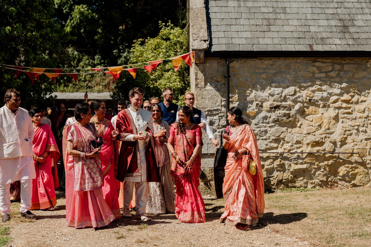 Wedding party in coral and pink dresses pose against stone wall exterior of rustic building.