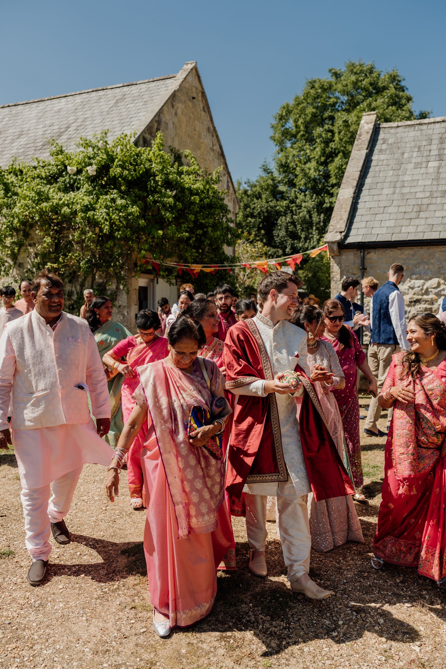 Wedding guests in pink and red attire socialize outdoors near stone buildings during a celebration.