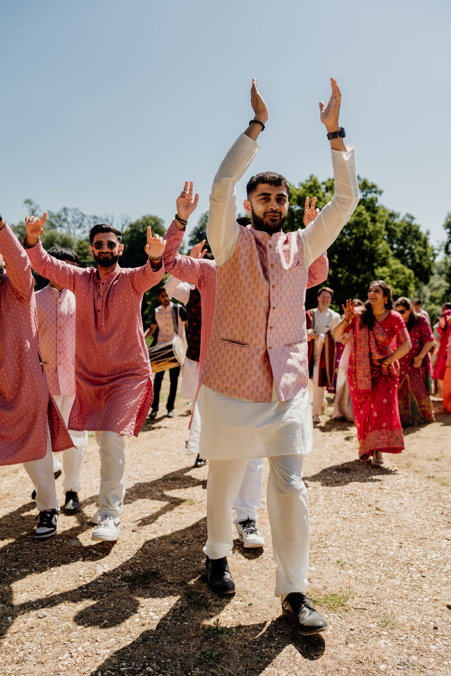 A group of wedding guests in coordinated pink and white outfits raise their arms joyfully while dancing outdoors.