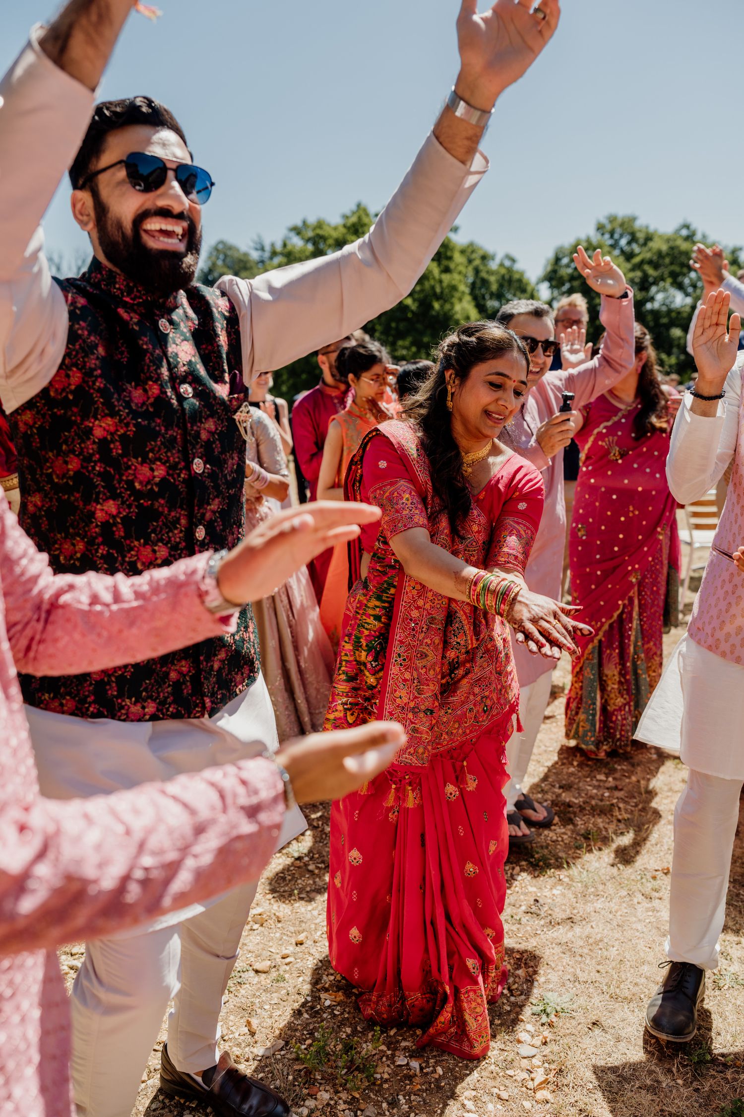 Wedding guests in coordinating red and pink outfits dance joyfully together outdoors on a sunny day.