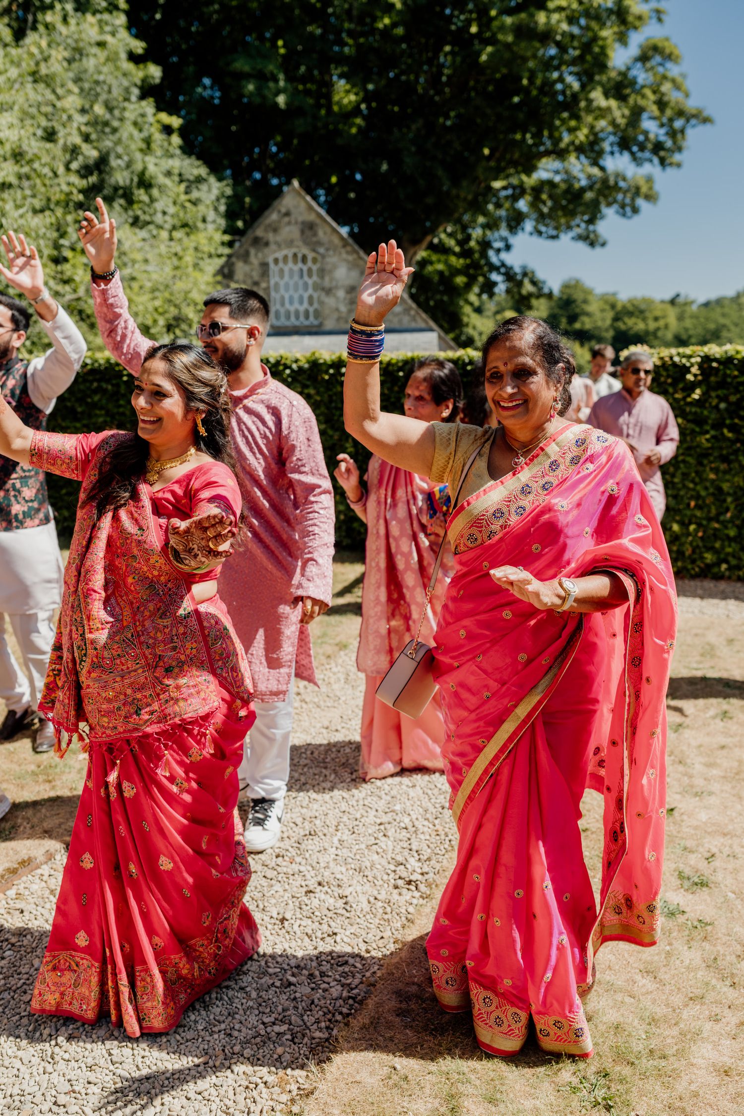 People in bright red and pink traditional outfits dance together on a sunny outdoor patio.