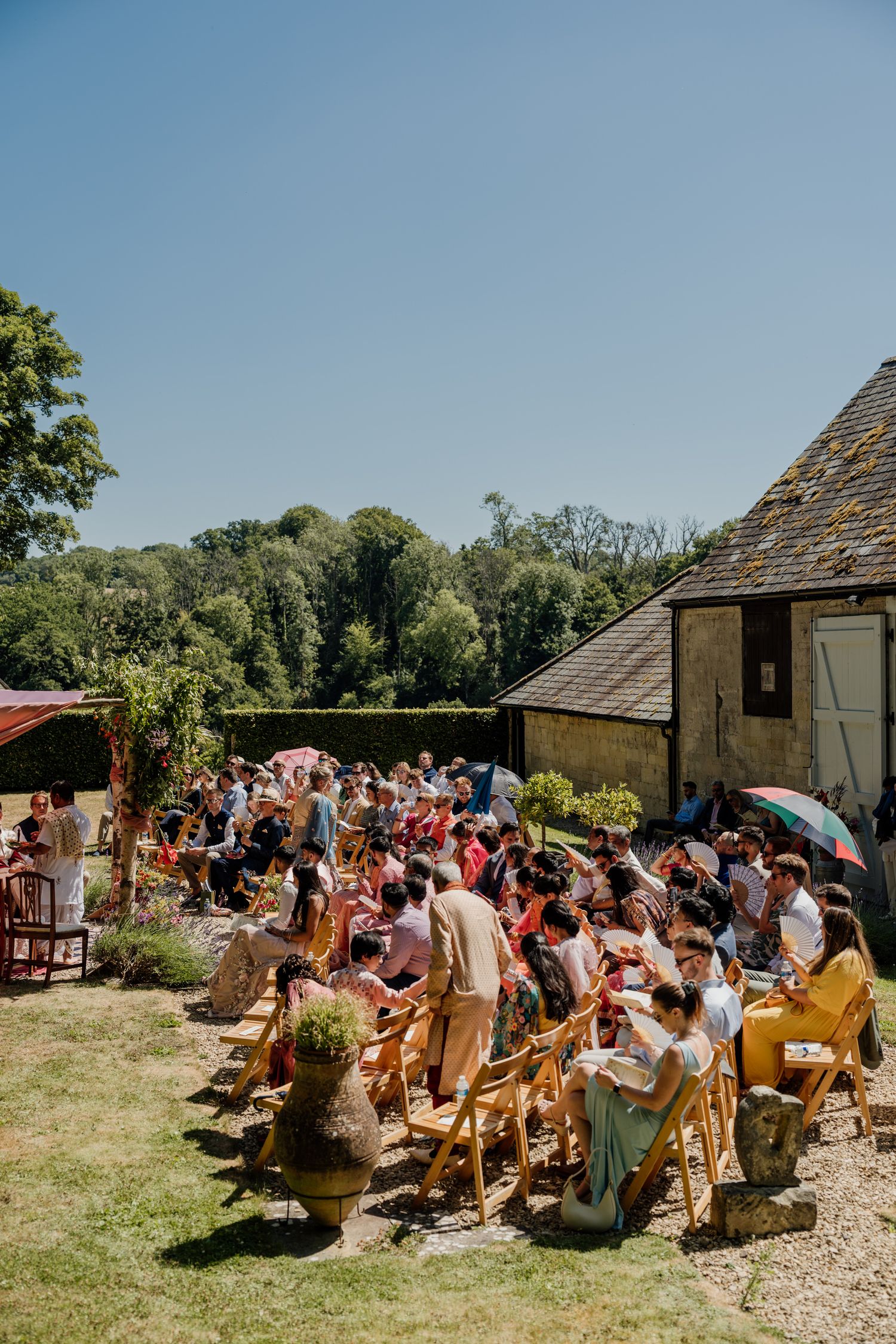 Large group of wedding guests seated outdoors at a rustic venue on a sunny summer day.