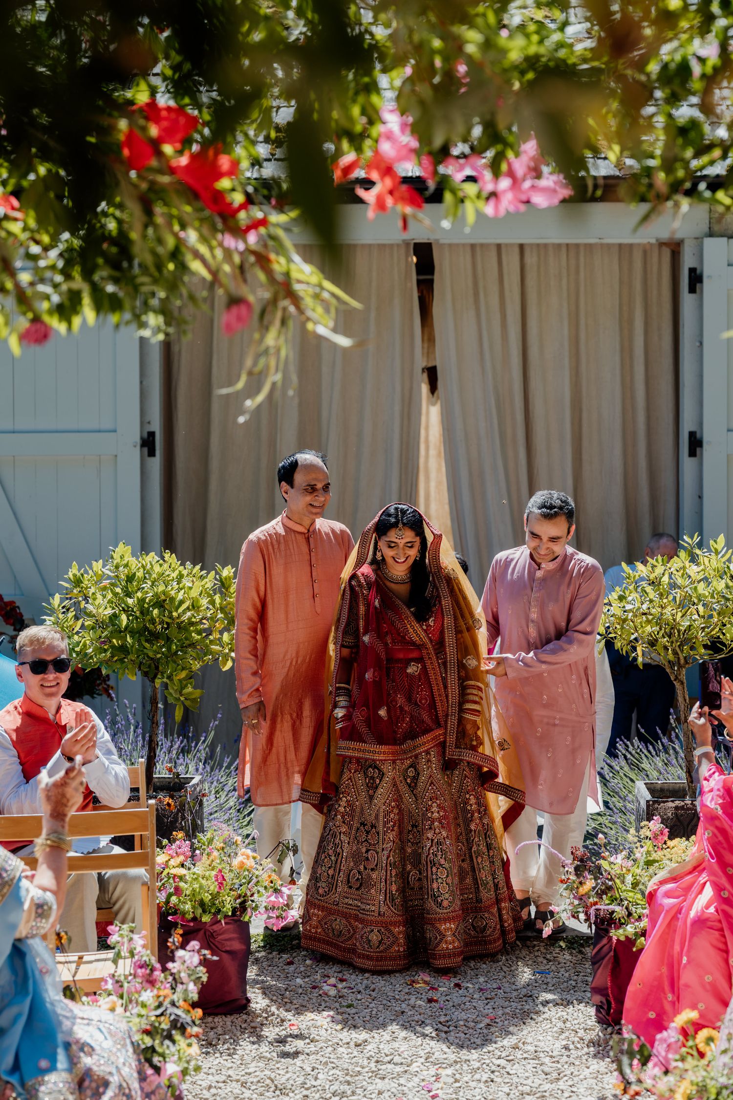 Guests in traditional Indian wedding attire gather outdoors under decorated trees with pink and red flowers.
