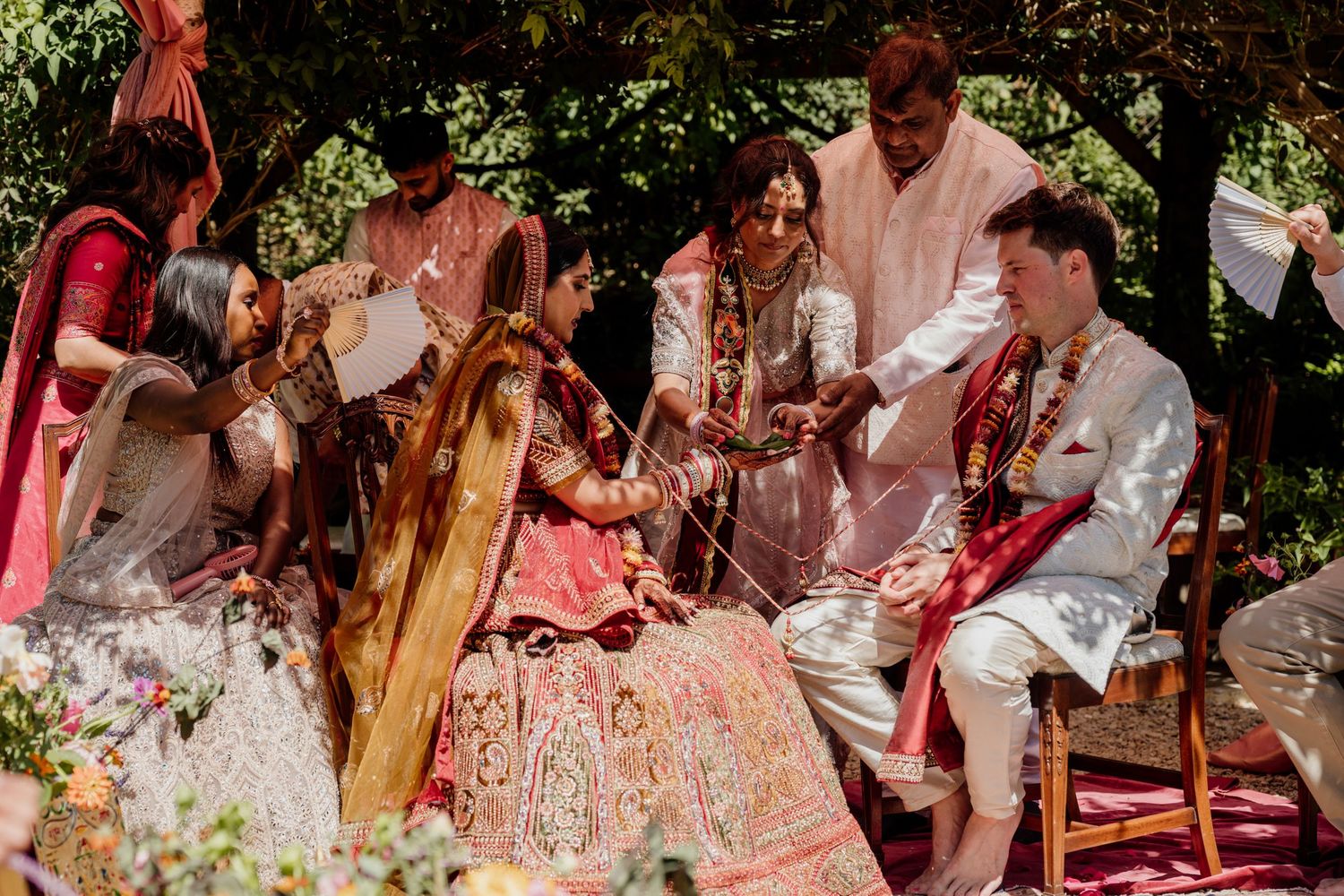 Couple in traditional Indian wedding attire performs ceremony rituals outdoors with musicians and guests in attendance.