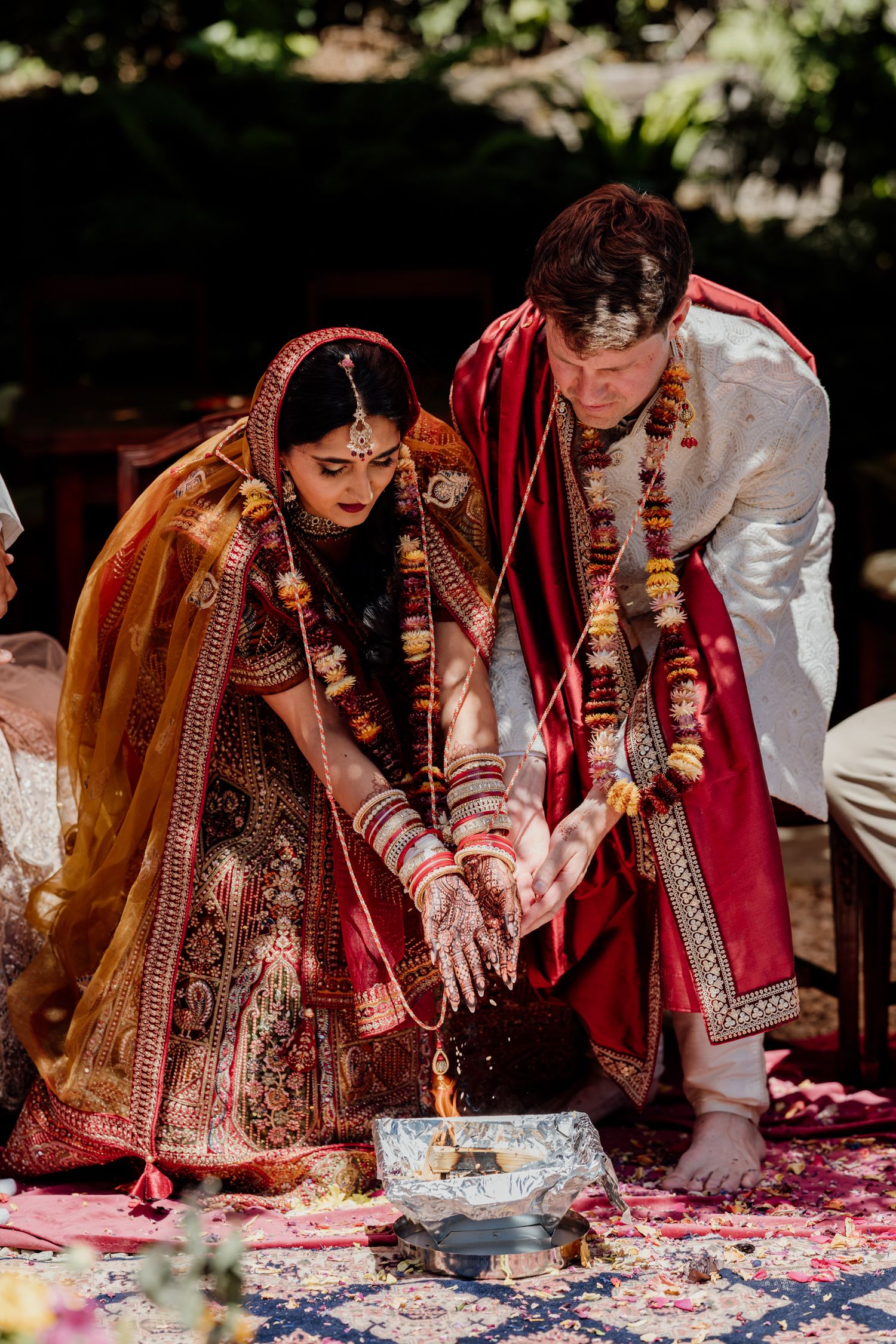 A couple performs a traditional Indian wedding ritual with rose petals during an outdoor ceremony.
