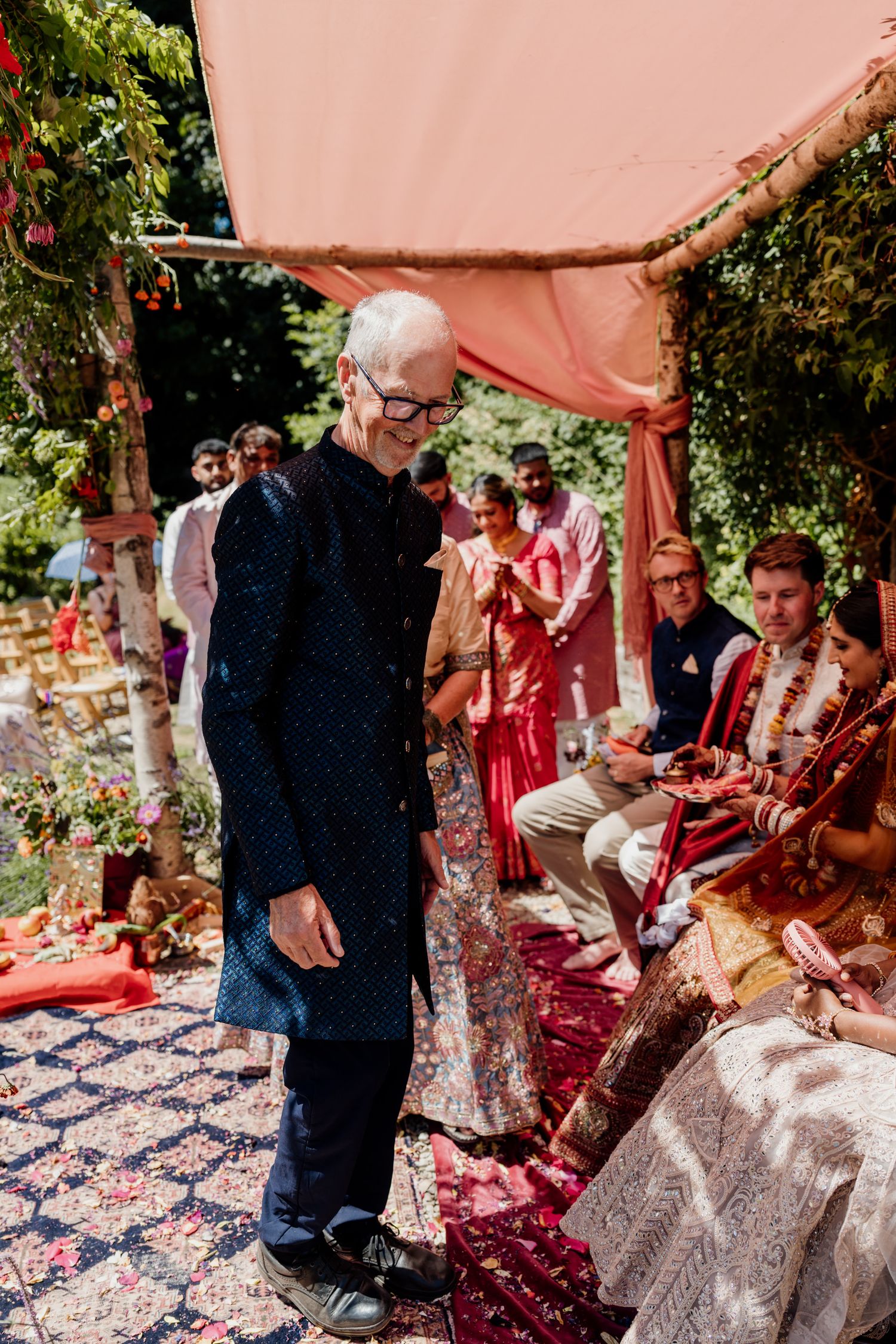Outdoor wedding ceremony under a pink canopy with guests seated on decorative rugs and cushions.