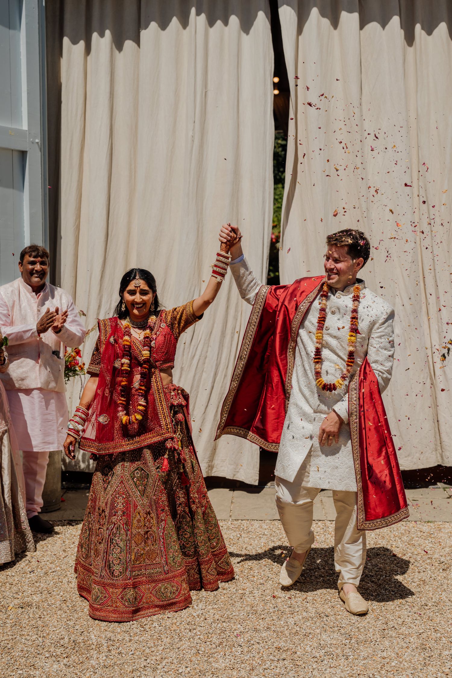 Wedding guests laughing and throwing red flower petals during a joyous celebration outdoors.