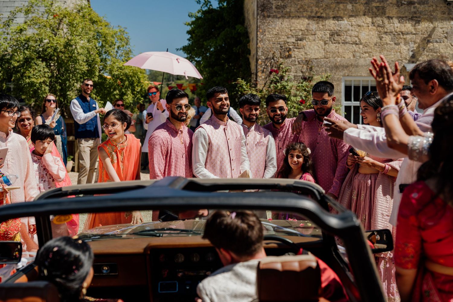 Wedding guests in pink and coral attire gather around a black luxury car during an outdoor celebration.