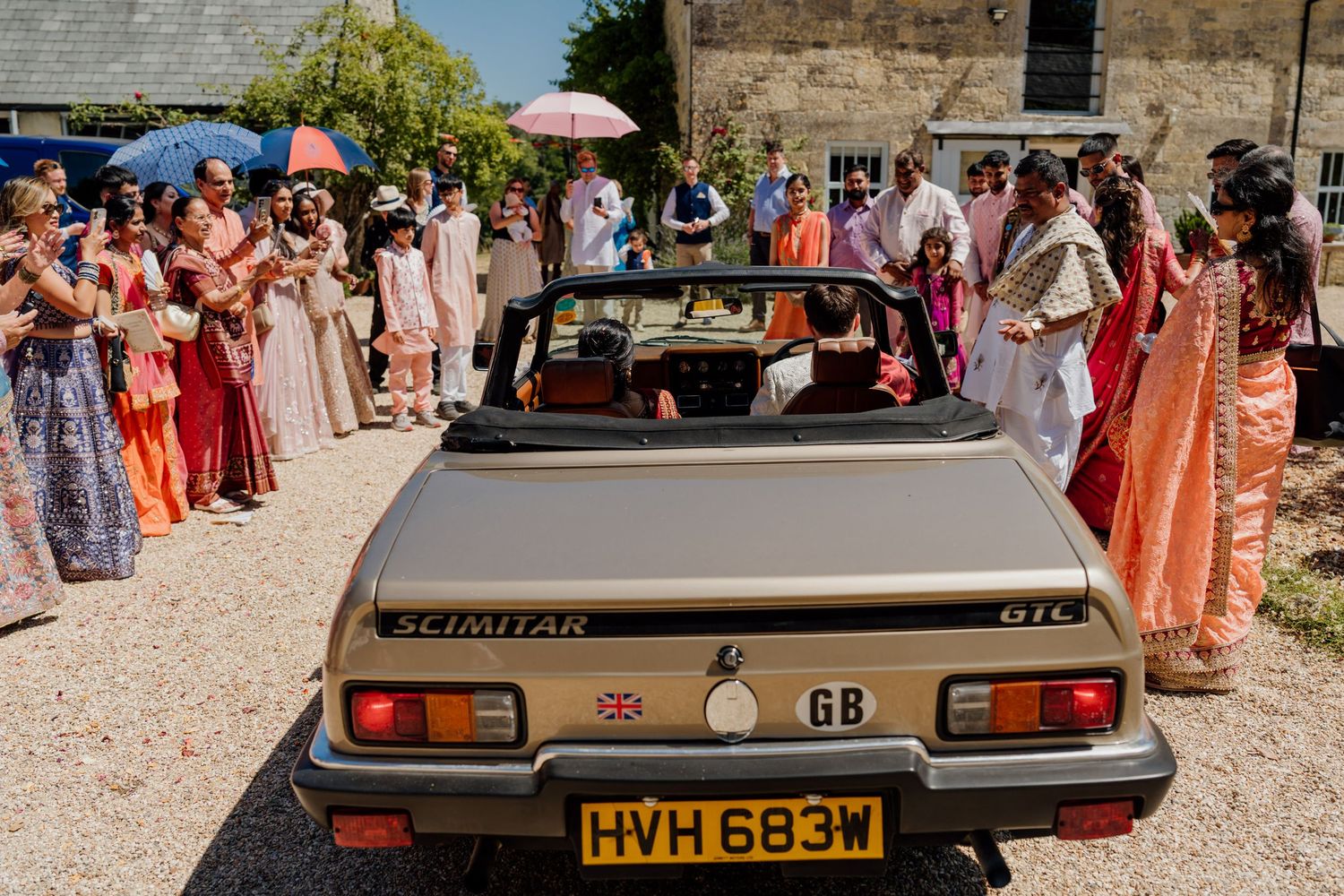 A classic convertible car is parked next to stone buildings during what appears to be an outdoor social gathering.