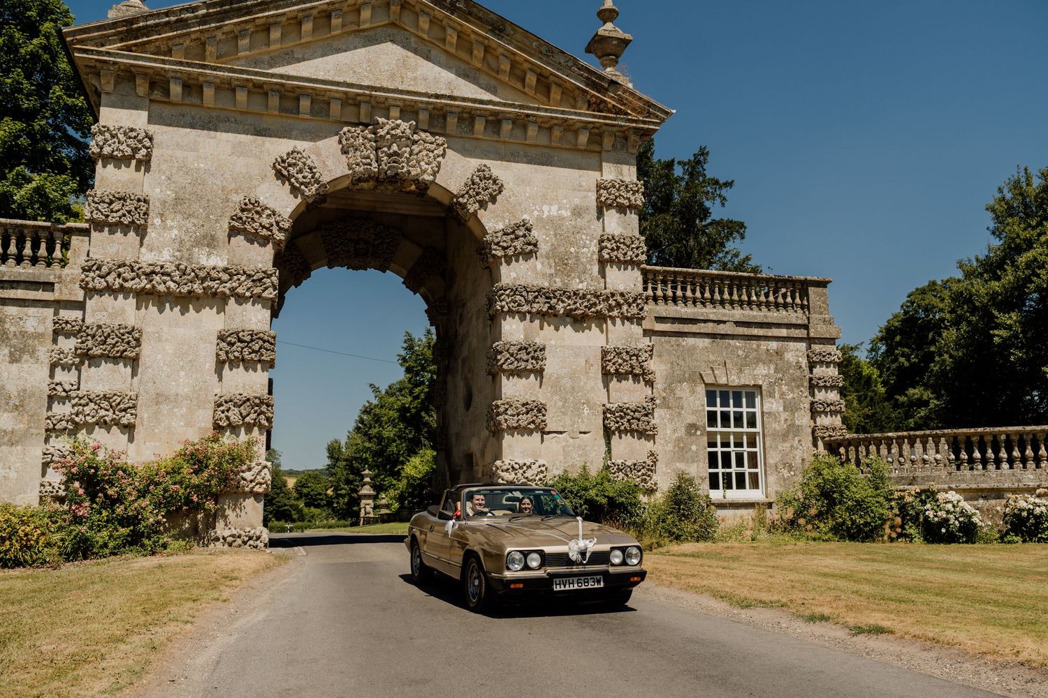 A vintage luxury car sits elegantly parked in front of a grand stone mansion on a sunny day.