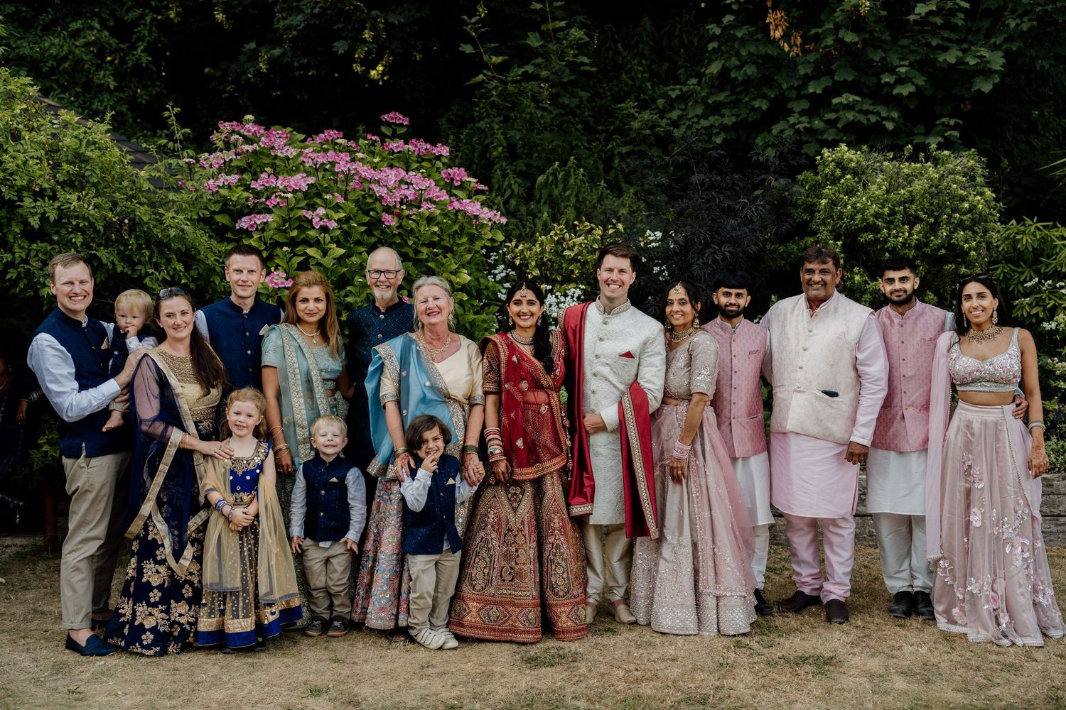 Large wedding group photo features guests in coordinated navy and pastel outfits in front of garden foliage.