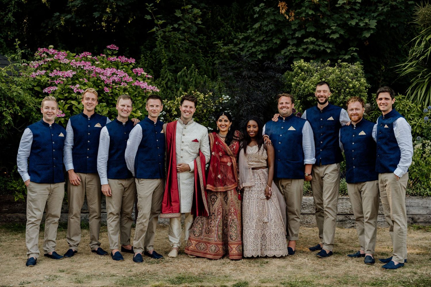 Wedding party lineup poses in casual attire with navy jackets against lush outdoor garden backdrop.