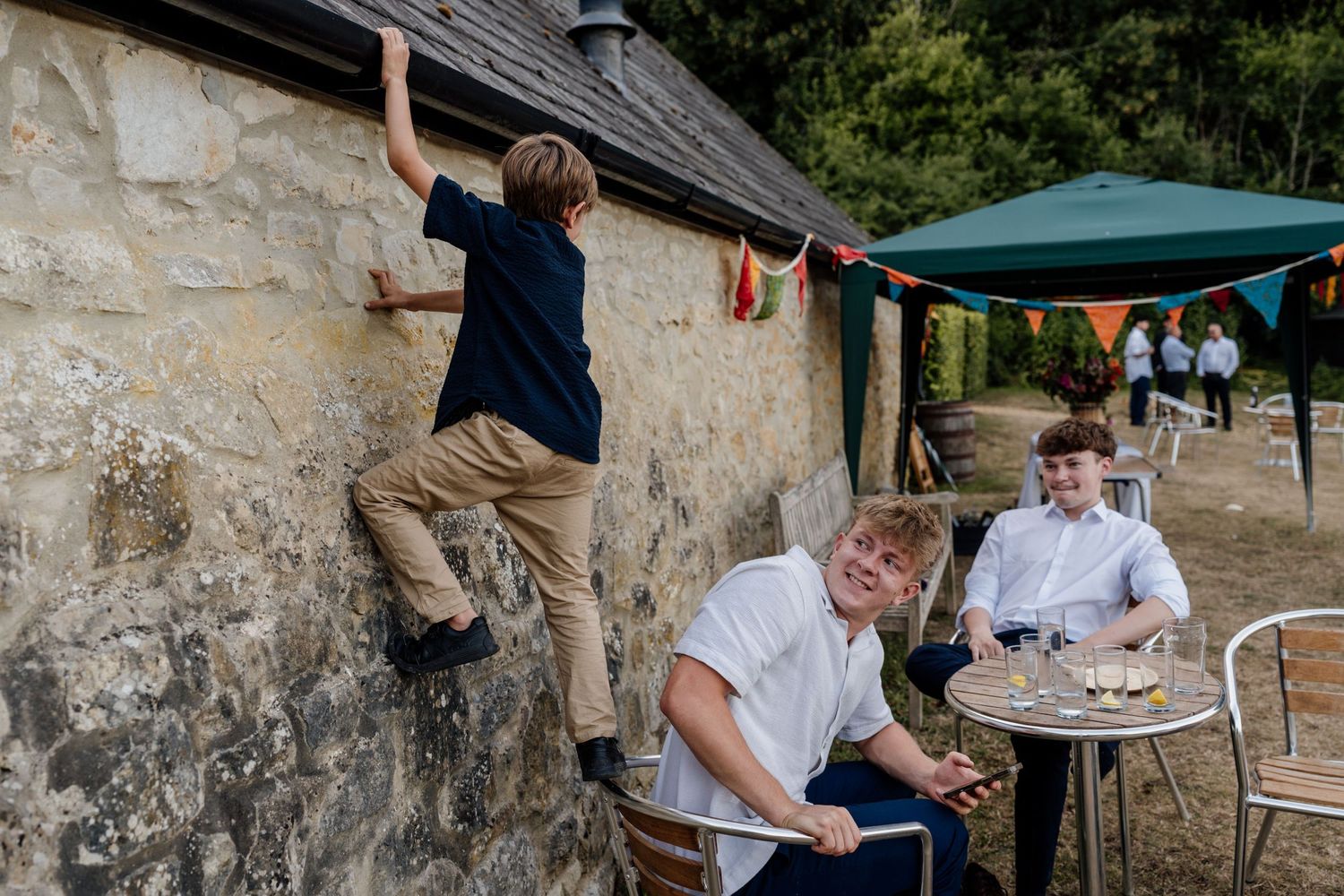 Outdoor casual gathering with people seated at tables under stone walls and green canopies.