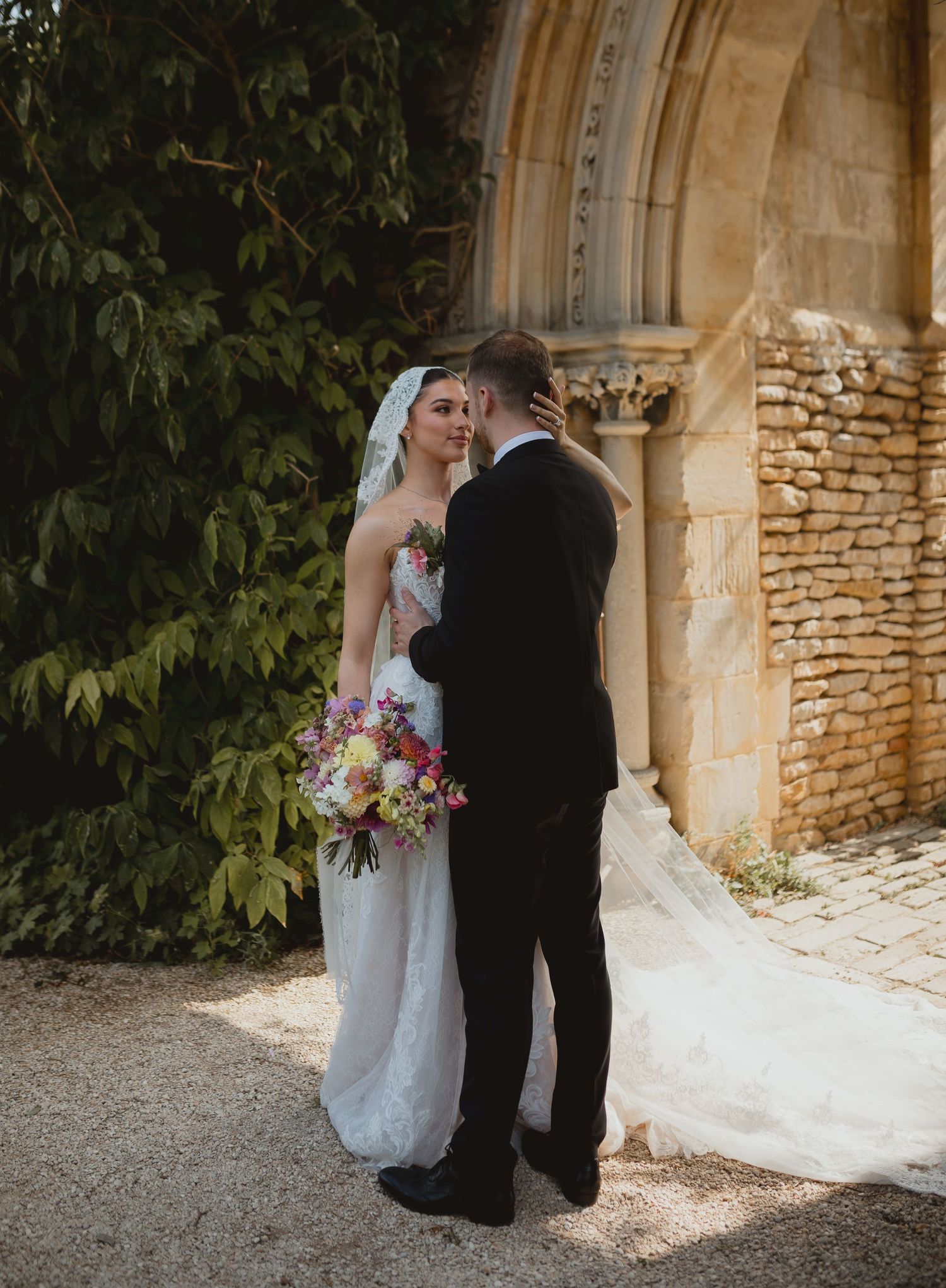 Romantic wedding couple poses near stone wall archway with bride holding vibrant floral bouquet in classic garden setting.
