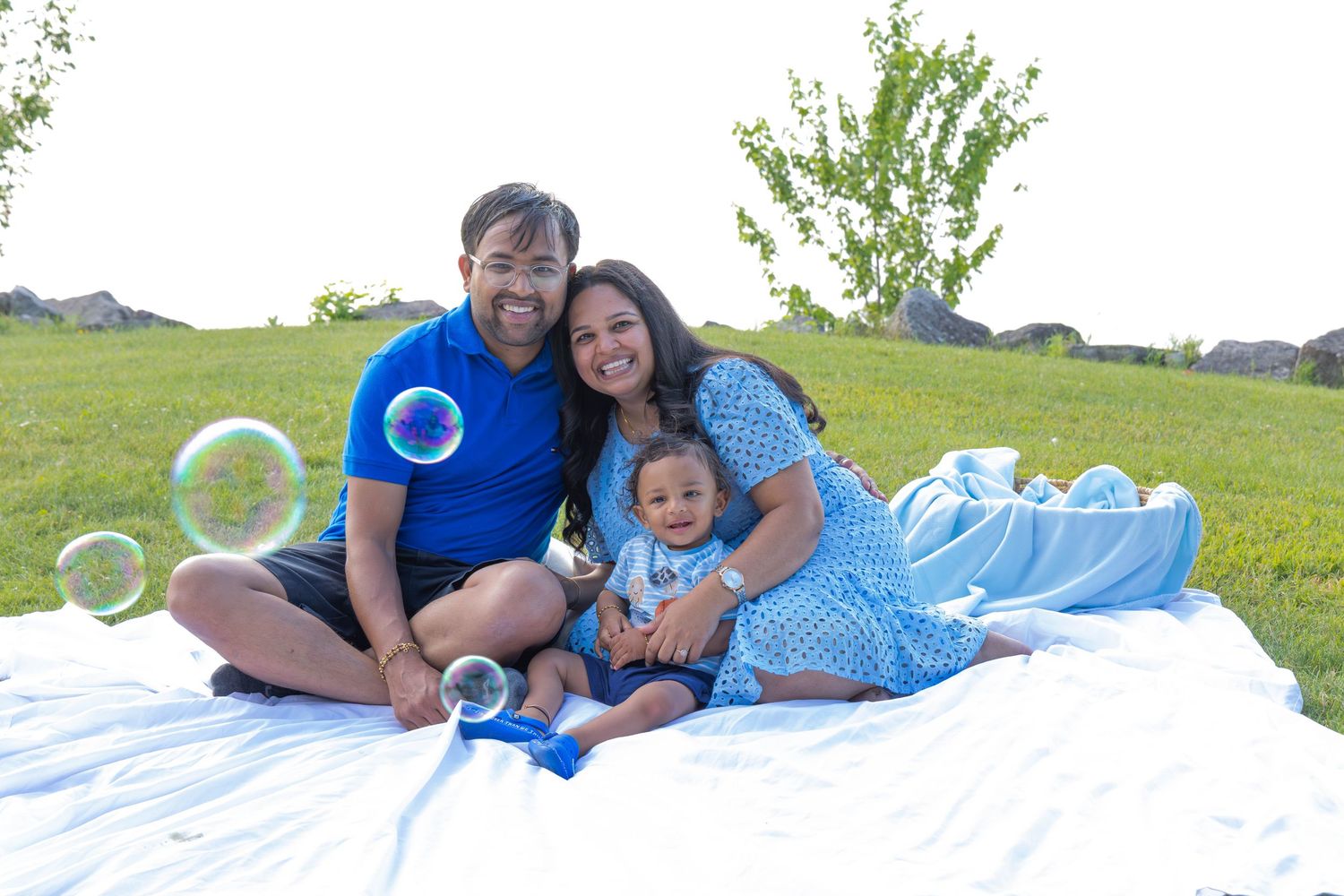 A family relaxes on a white blanket in a grassy field with soap bubbles floating around them on a sunny day.